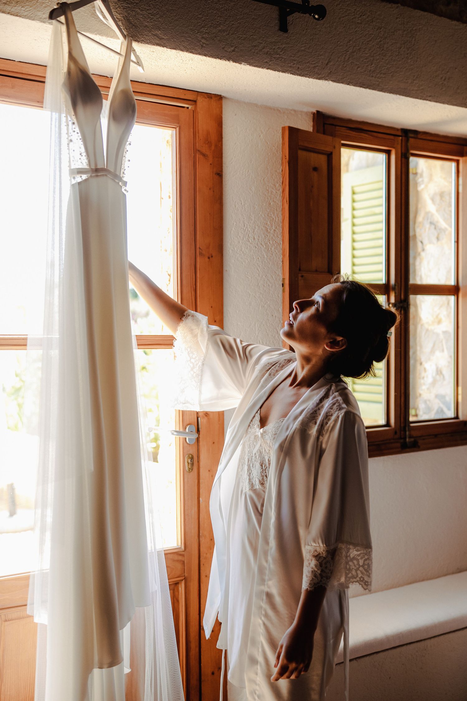 Bride in white lace robe reaching up to touch elegant wedding dress hanging by sunlit window.