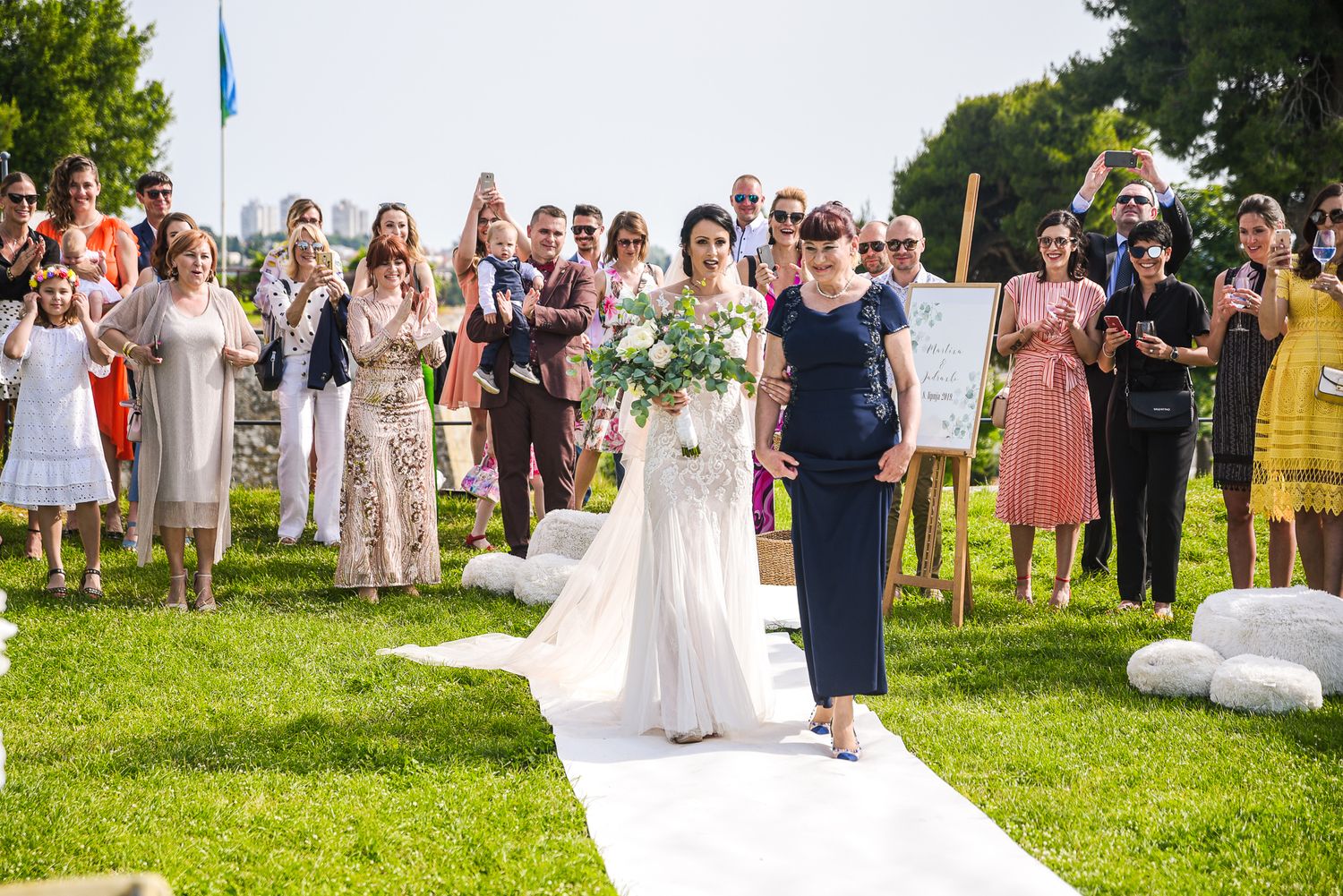 Bride in white gown walks down aisle with escort at sunny outdoor wedding ceremony.