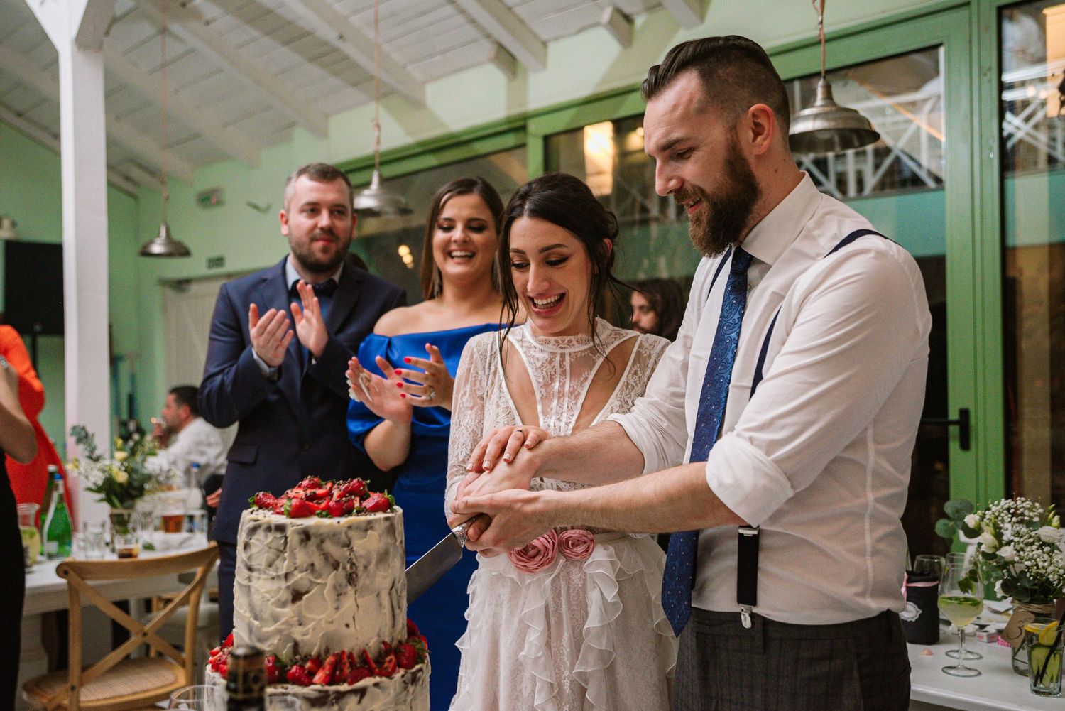 Bride and groom cut their two-tier strawberry wedding cake while guests applaud.