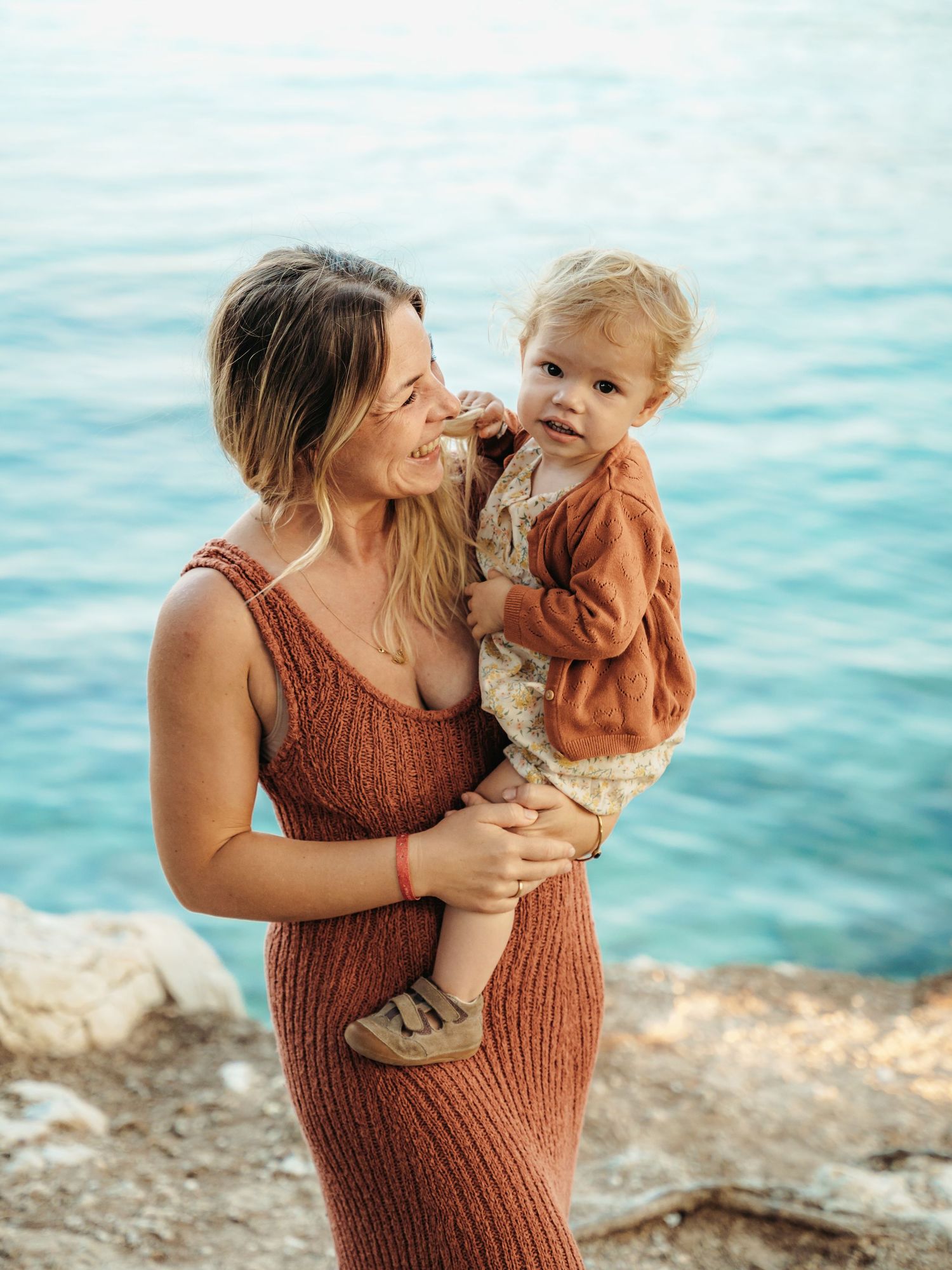 A smiling mother in a rust knit dress holds her toddler by the turquoise sea.
