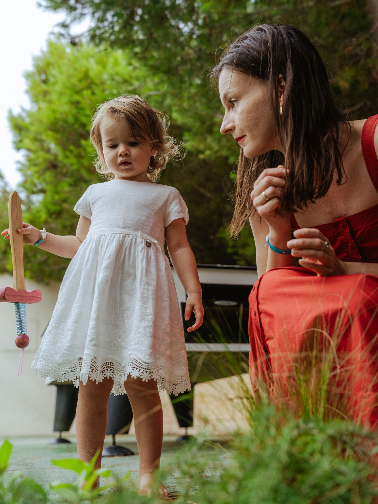 A woman in a red dress watches a toddler girl in white dress playing with a wooden toy outside.