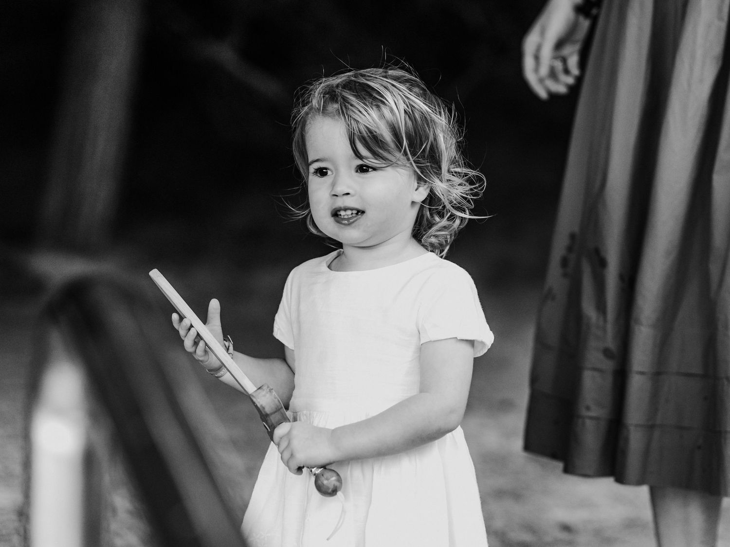 Toddler girl in white dress holding a wooden toy outdoors in black and white photo.
