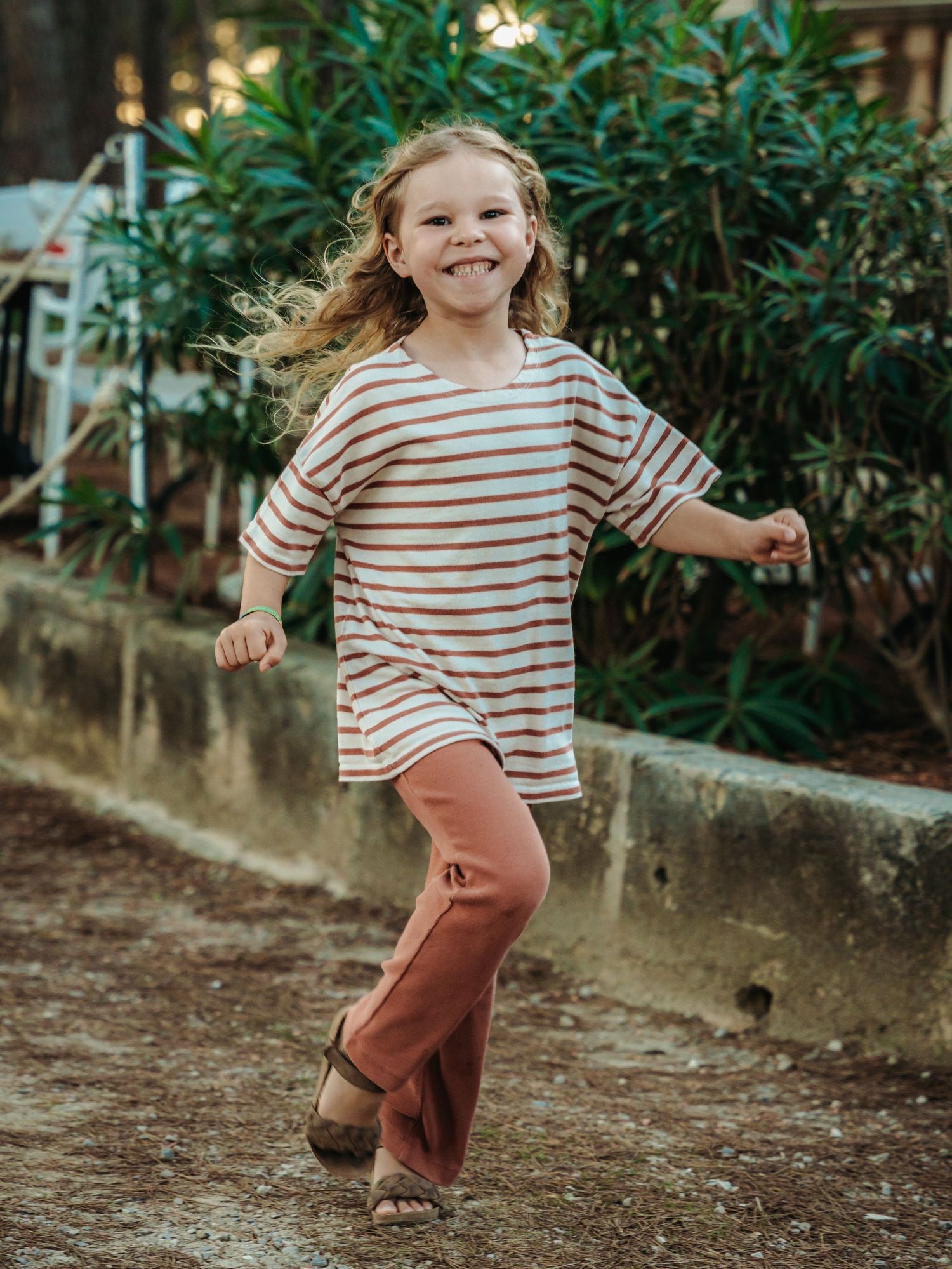 Happy young girl with curly hair running outdoors in a striped shirt and pink pants.