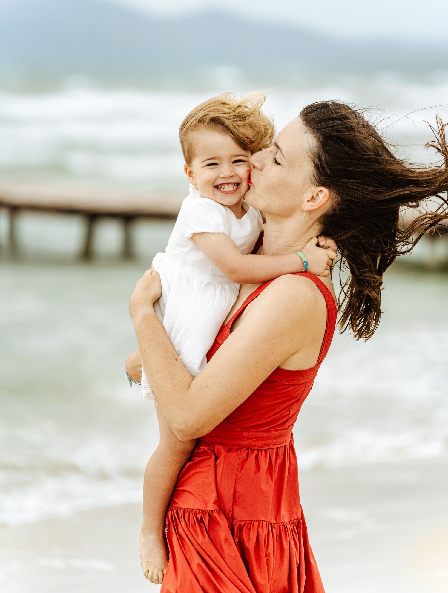 Mother in red dress kissing happy baby on windy beach with pier backdrop.