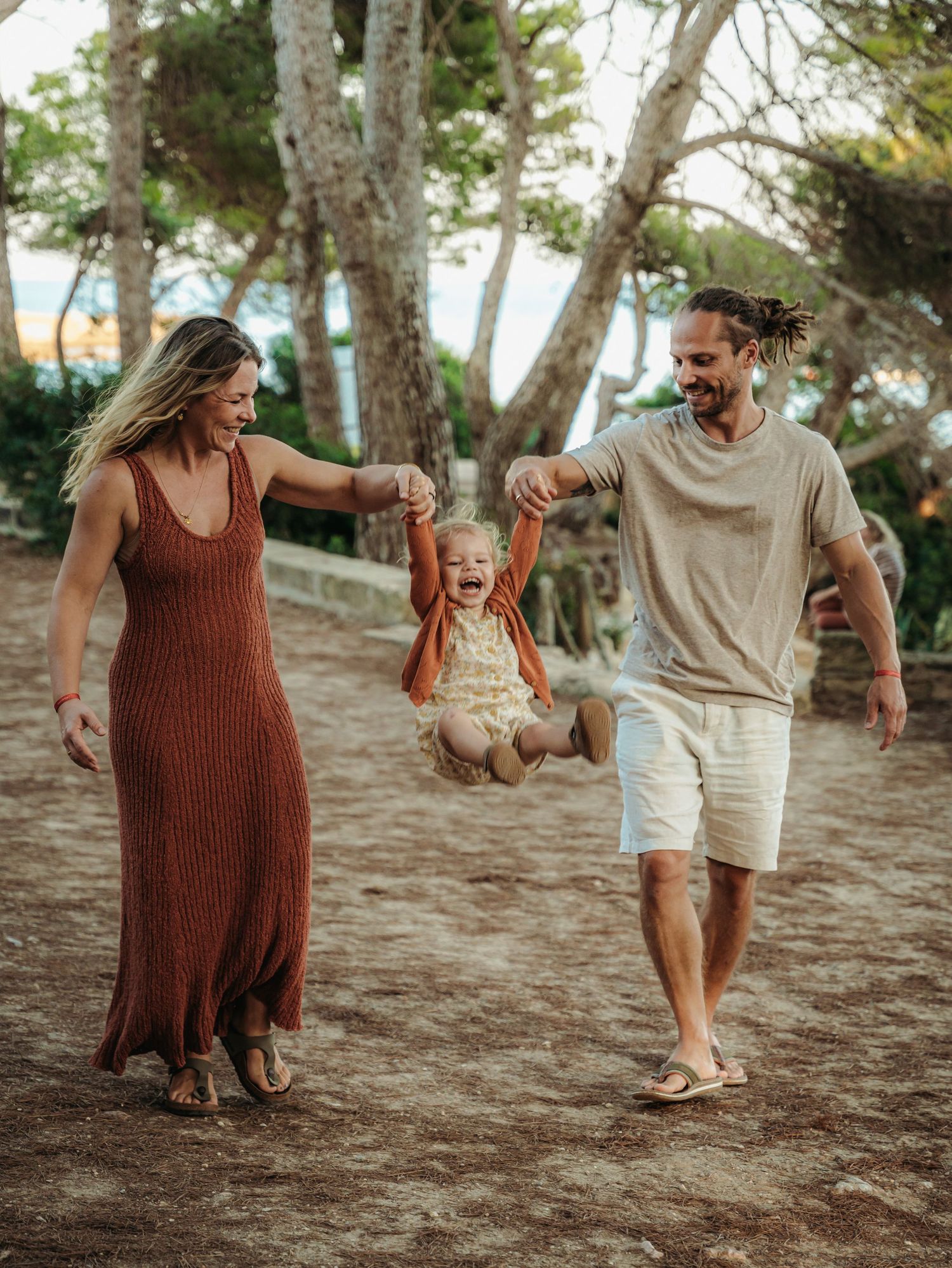 Smiling parents swinging laughing toddler between them on sandy ground among pine trees.