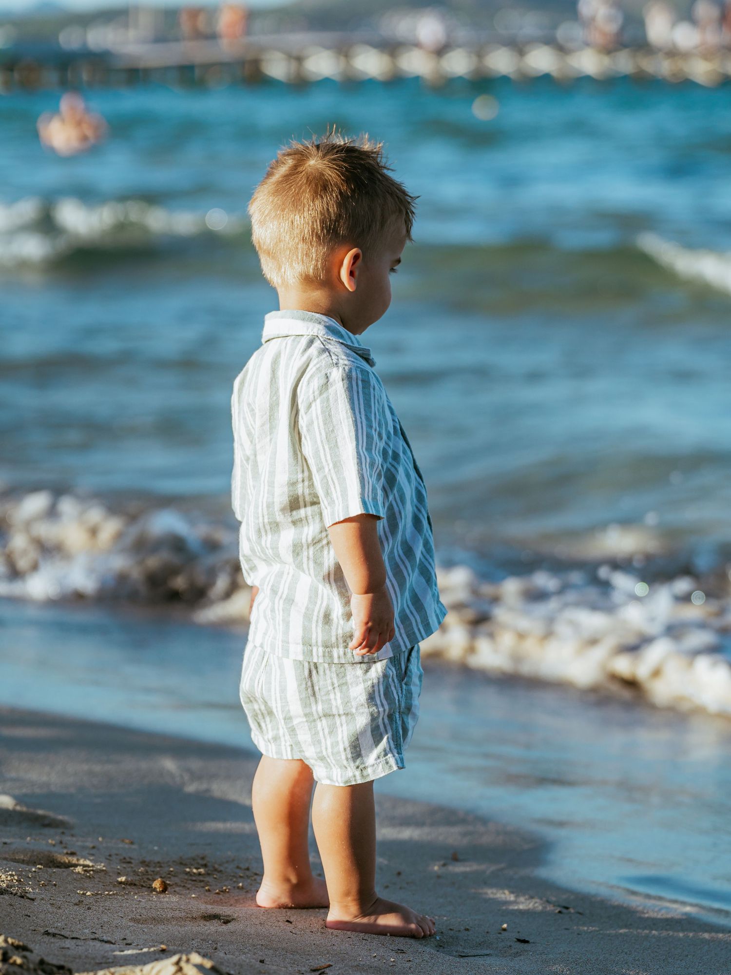 Toddler boy in striped outfit stands barefoot at water's edge gazing at ocean waves.