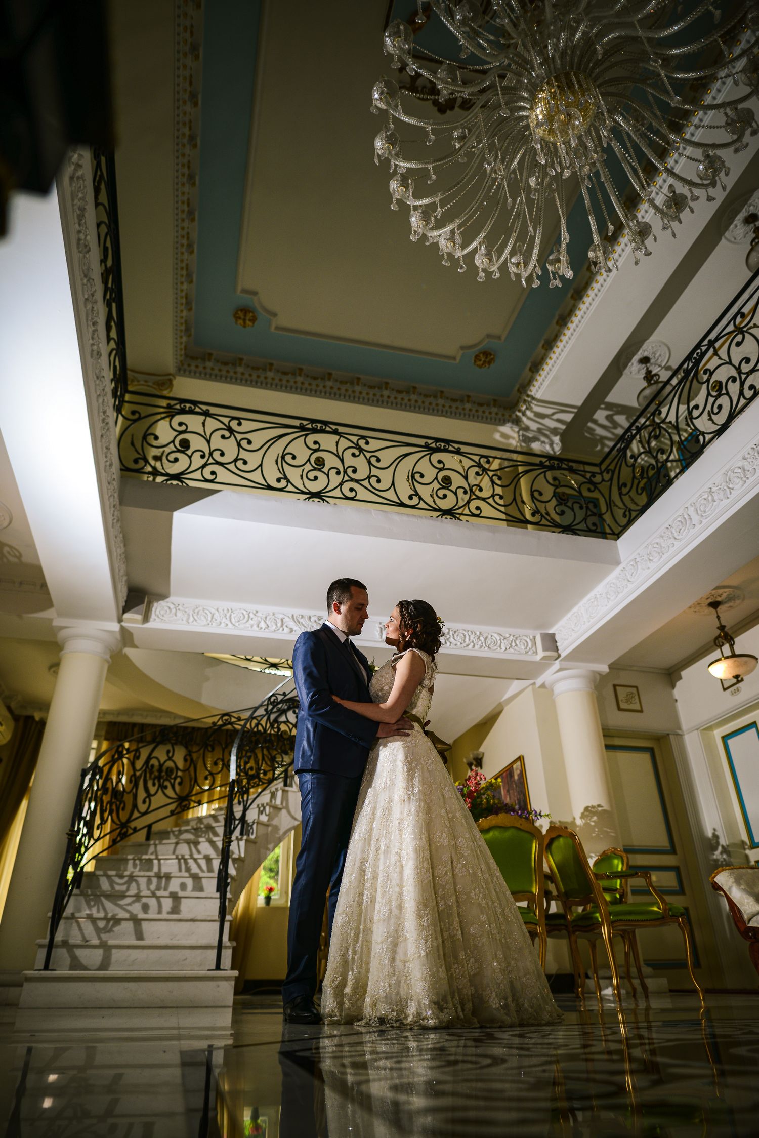 Elegant bride and groom embrace in a grand ornate hotel lobby beneath a crystal chandelier.
