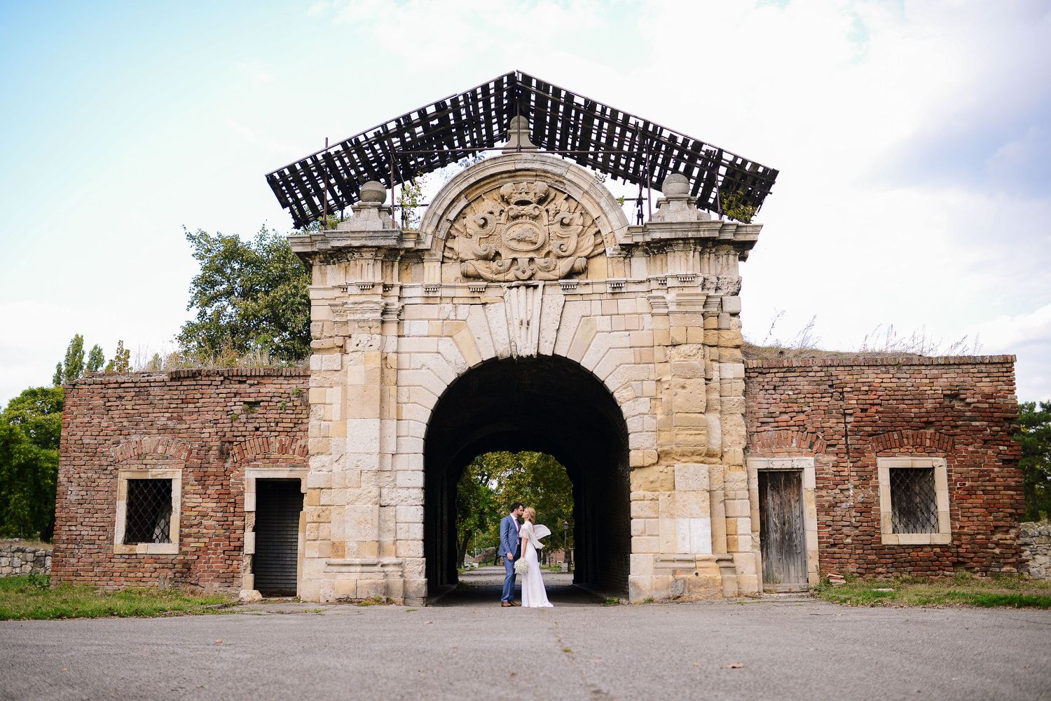 Bride and groom kiss beneath an ornate historic stone gate archway in a grand outdoor wedding portrait.