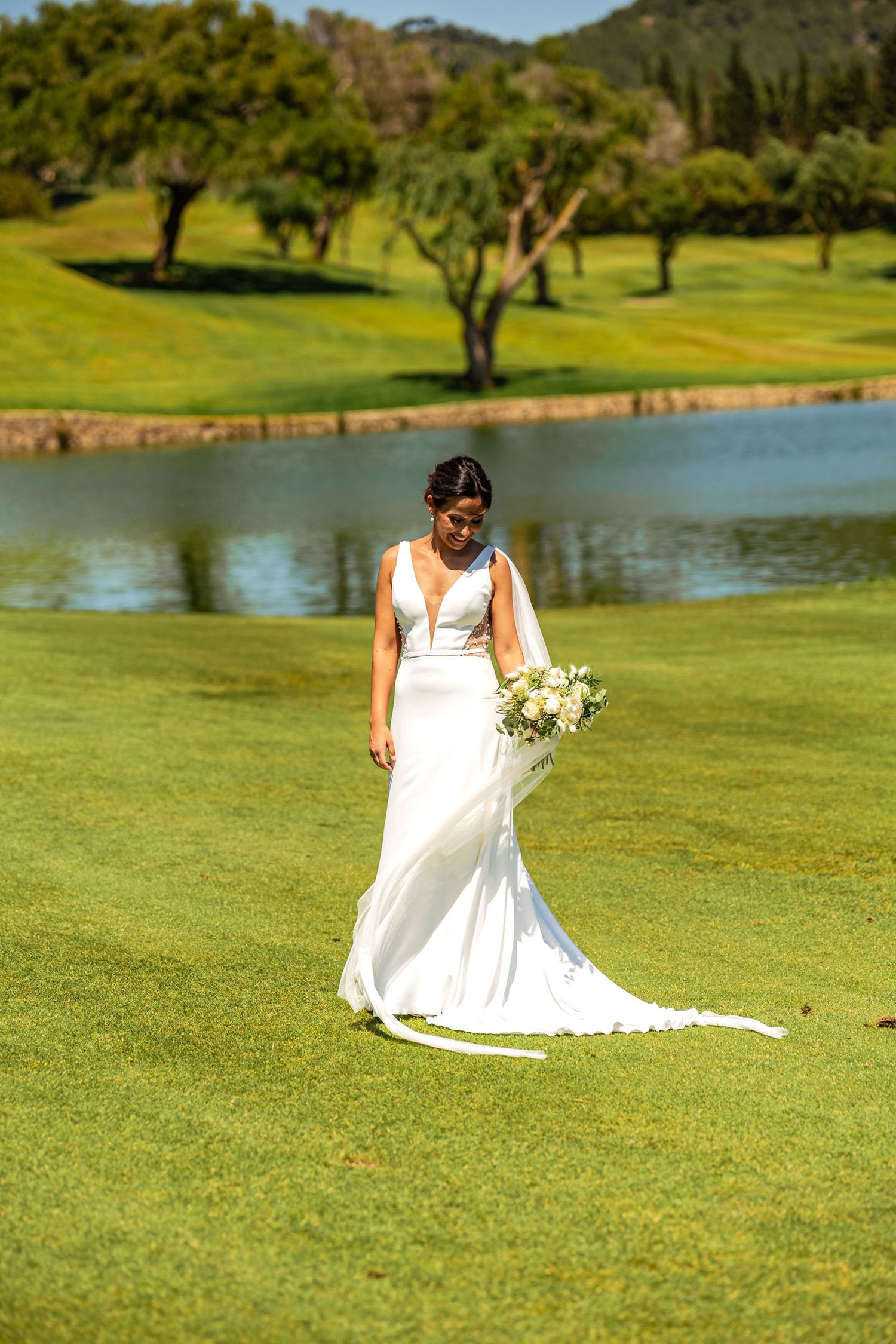 Elegant bride in white gown holding bouquet walking on lush green golf course by a pond.
