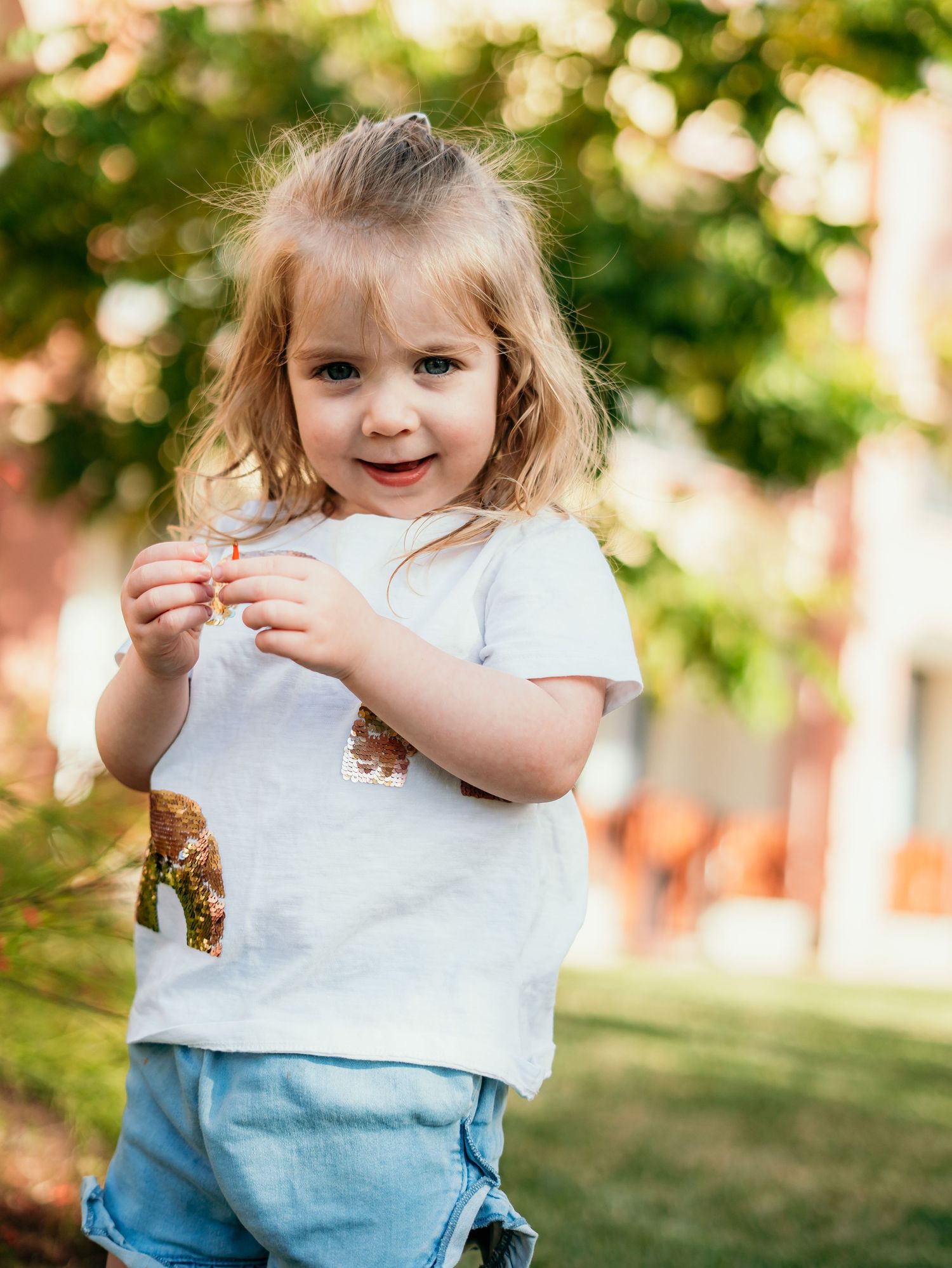 Smiling toddler girl in white t-shirt and denim shorts holding something small in a sunny garden.