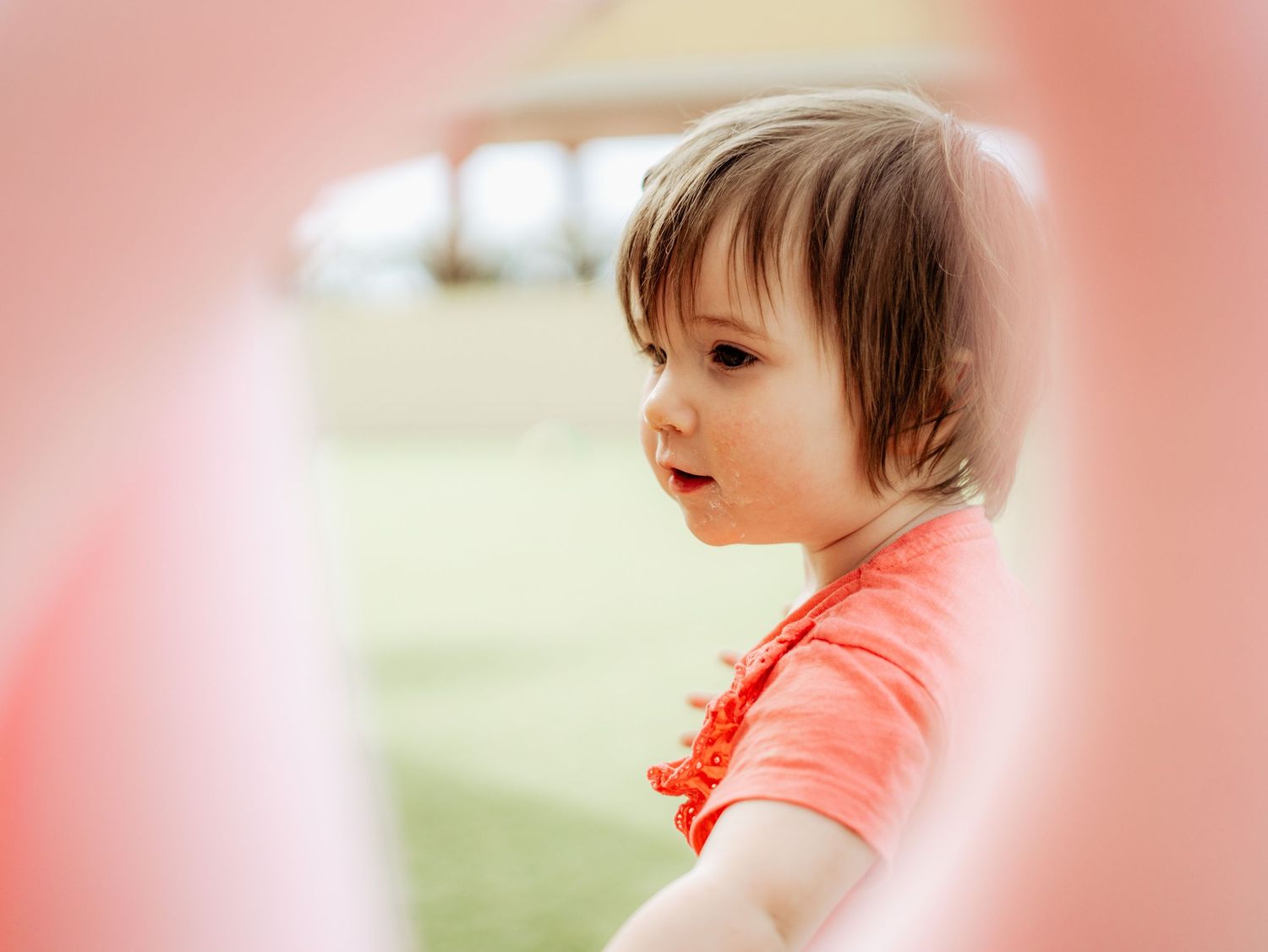 Close-up of a toddler girl in red top peeking through a pink pool noodle outdoors.