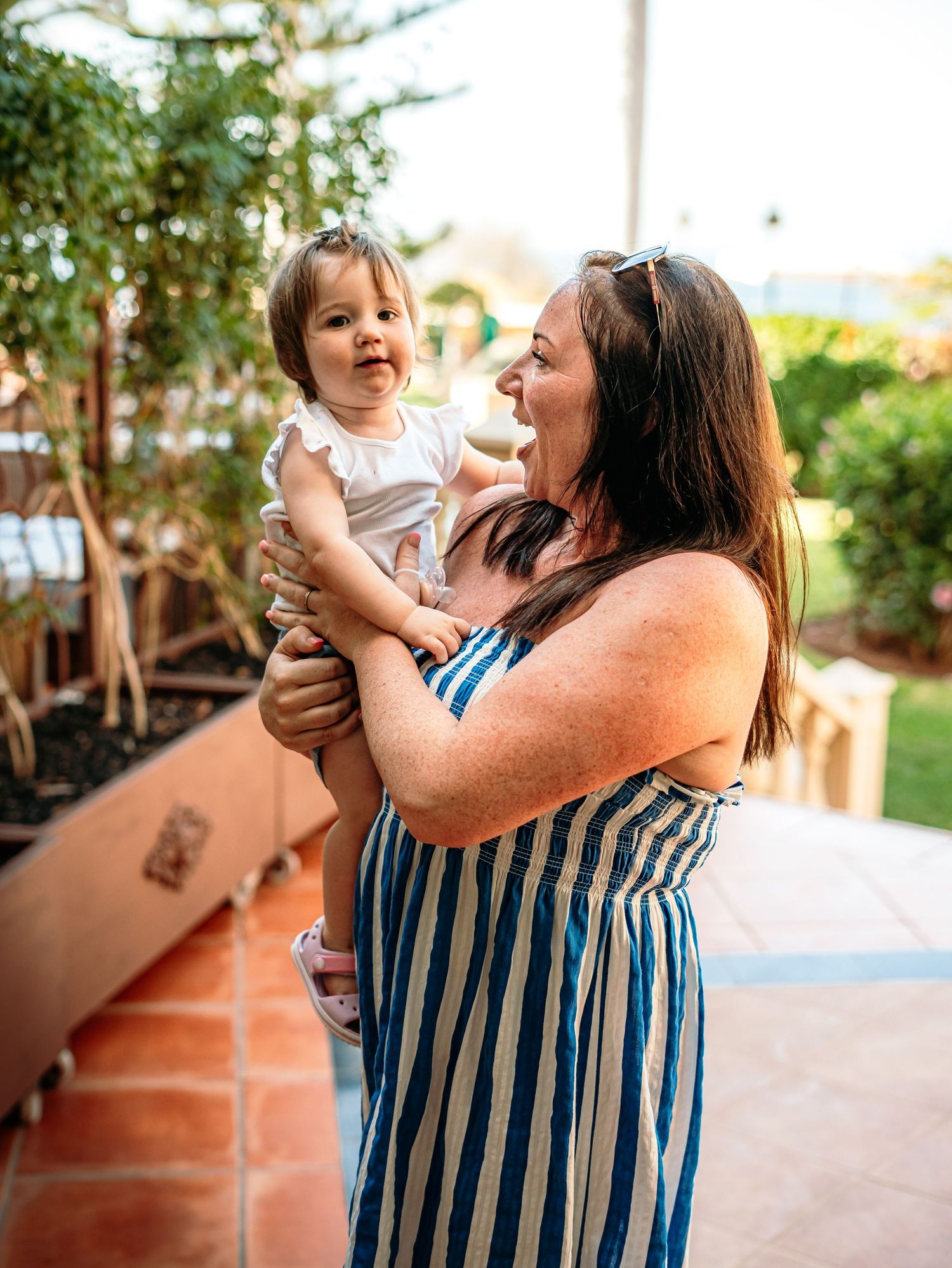 A woman in a striped dress holds a smiling baby girl outdoors on a sunny patio.