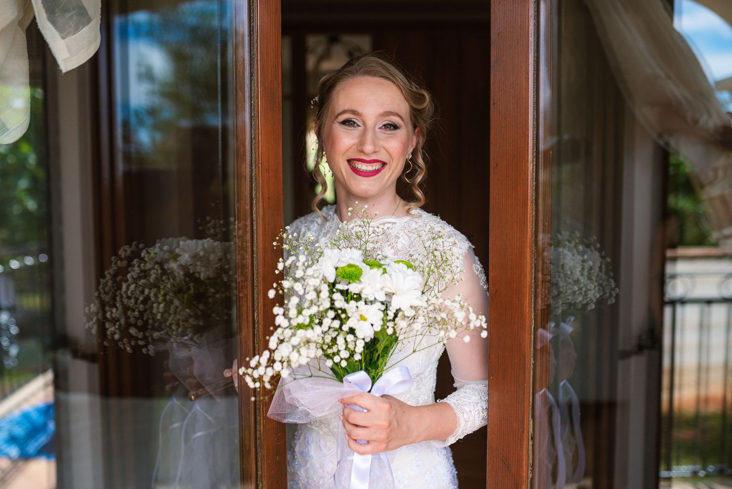 Smiling bride in lace gown holding white baby's breath bouquet in doorway.