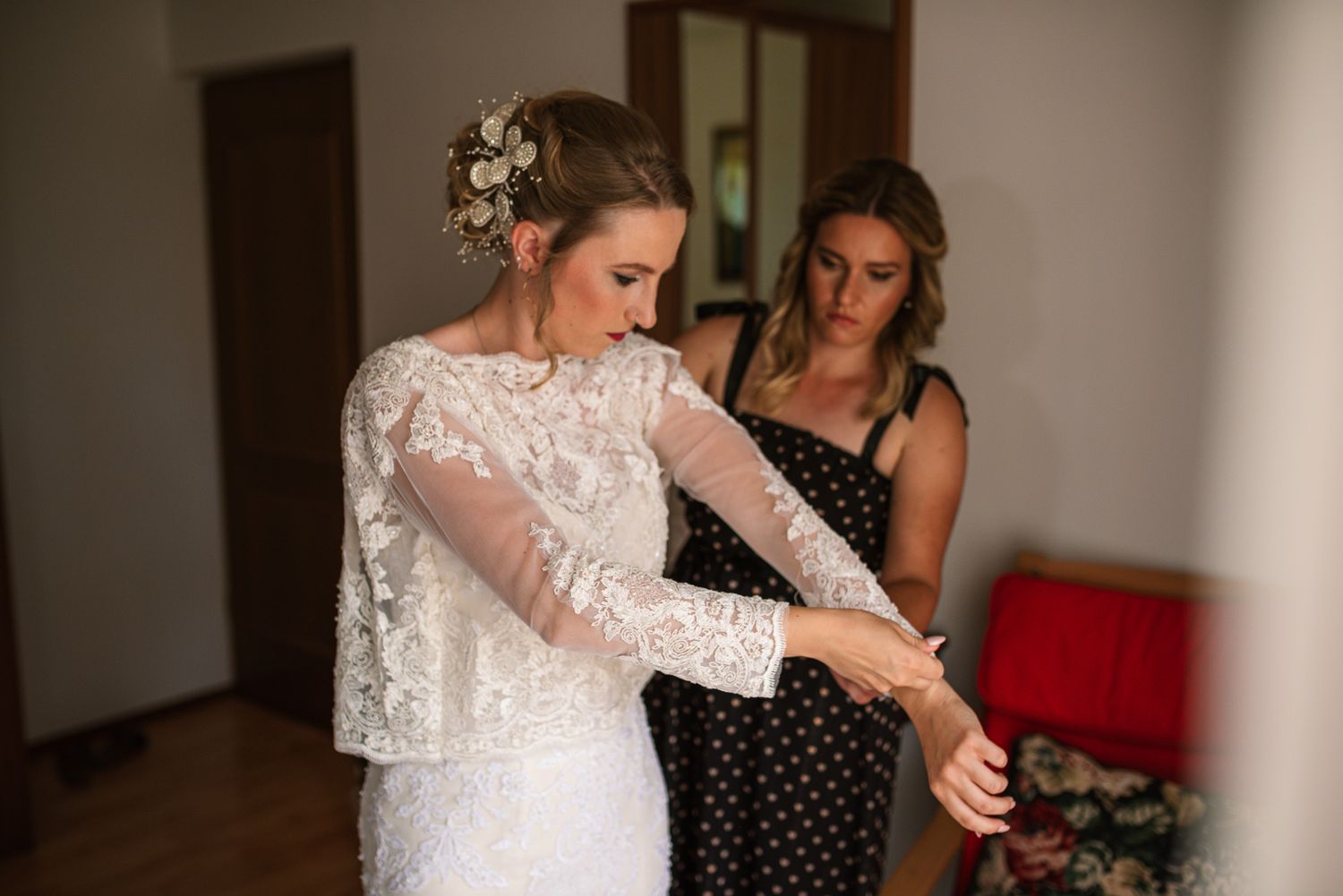 Bride in lace wedding dress getting help from bridesmaid in black polka dot dress during wedding preparations.