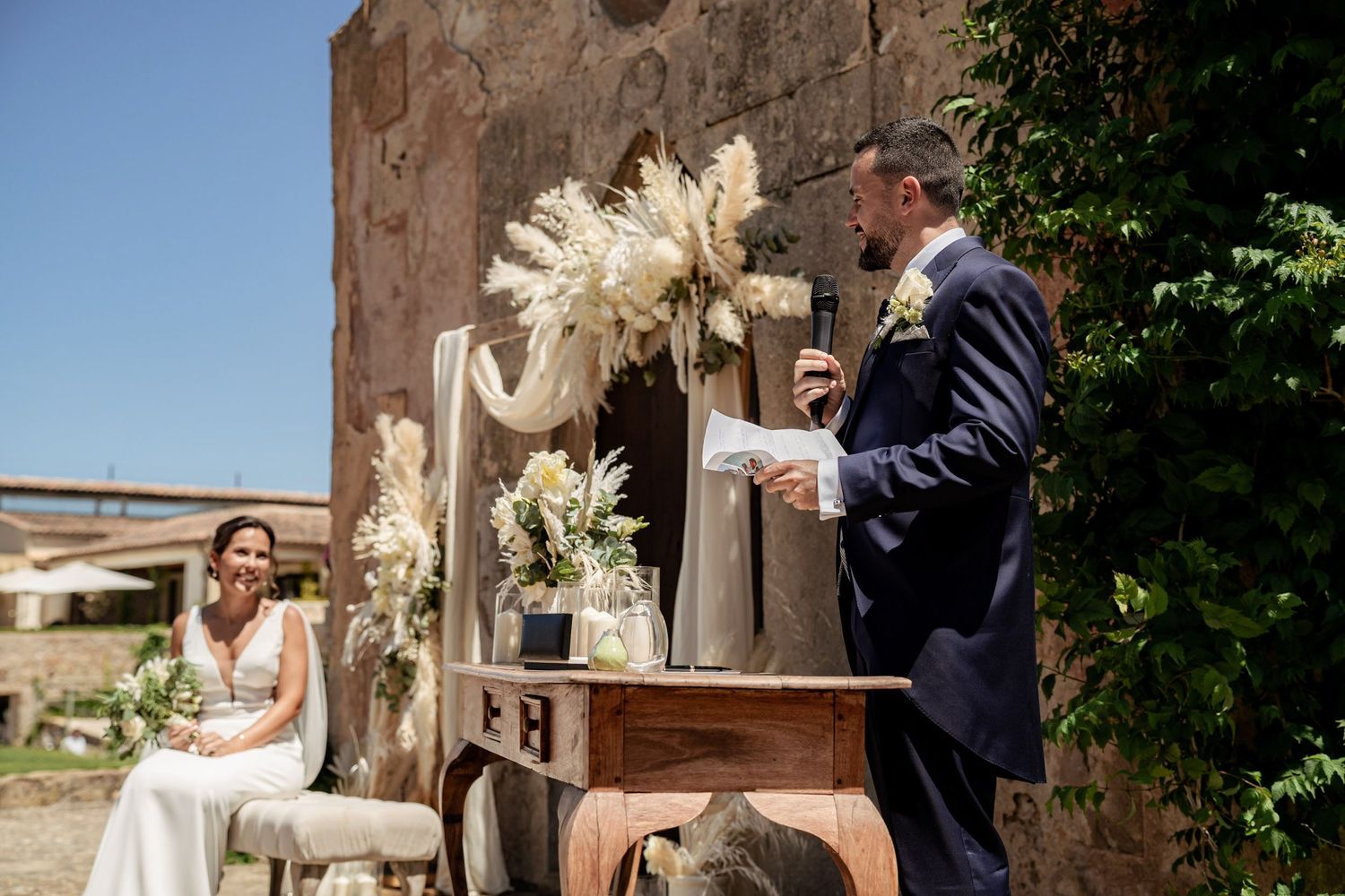 Groom reads vows at rustic wooden podium as bride smiles during sunny outdoor wedding ceremony.