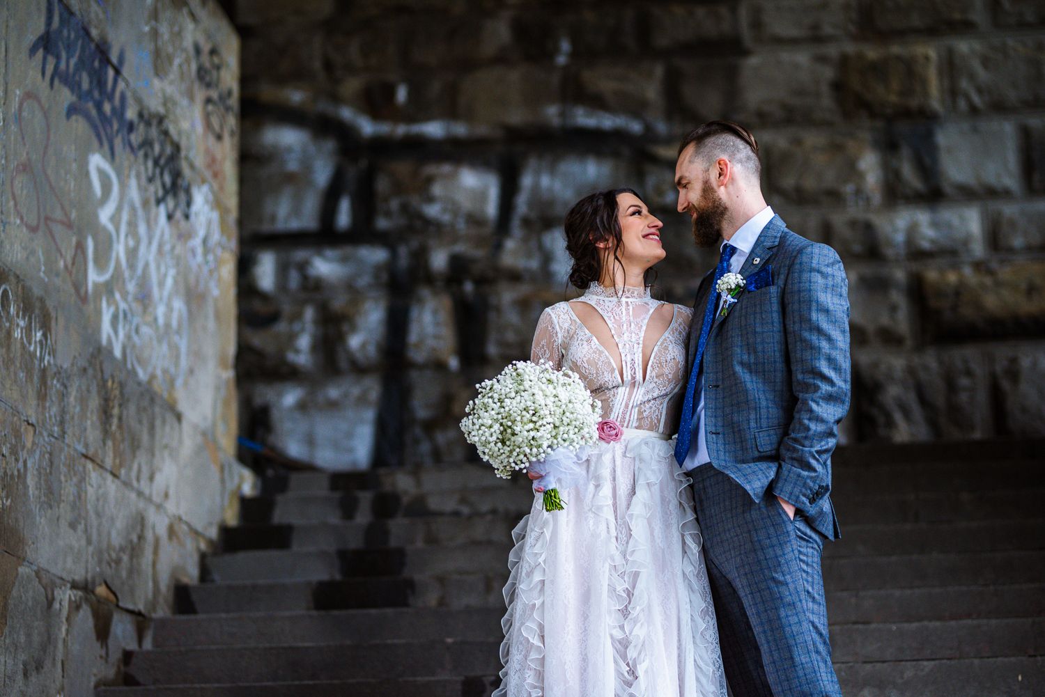 Bride and groom sharing an intimate moment on stone steps beside a graffiti-covered wall.