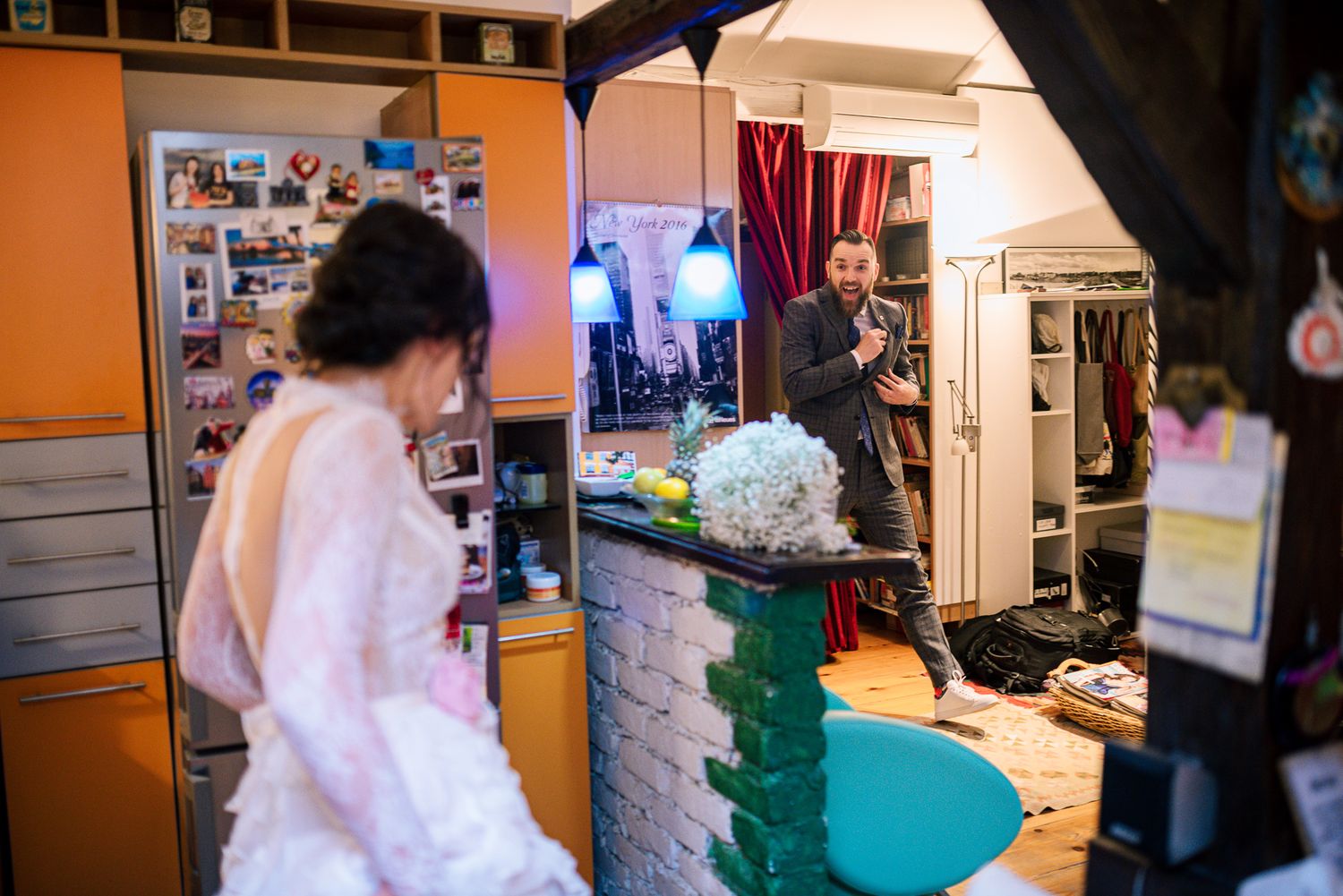 Bride in white lace dress getting ready in a decorated room while a man in a suit watches from the background.