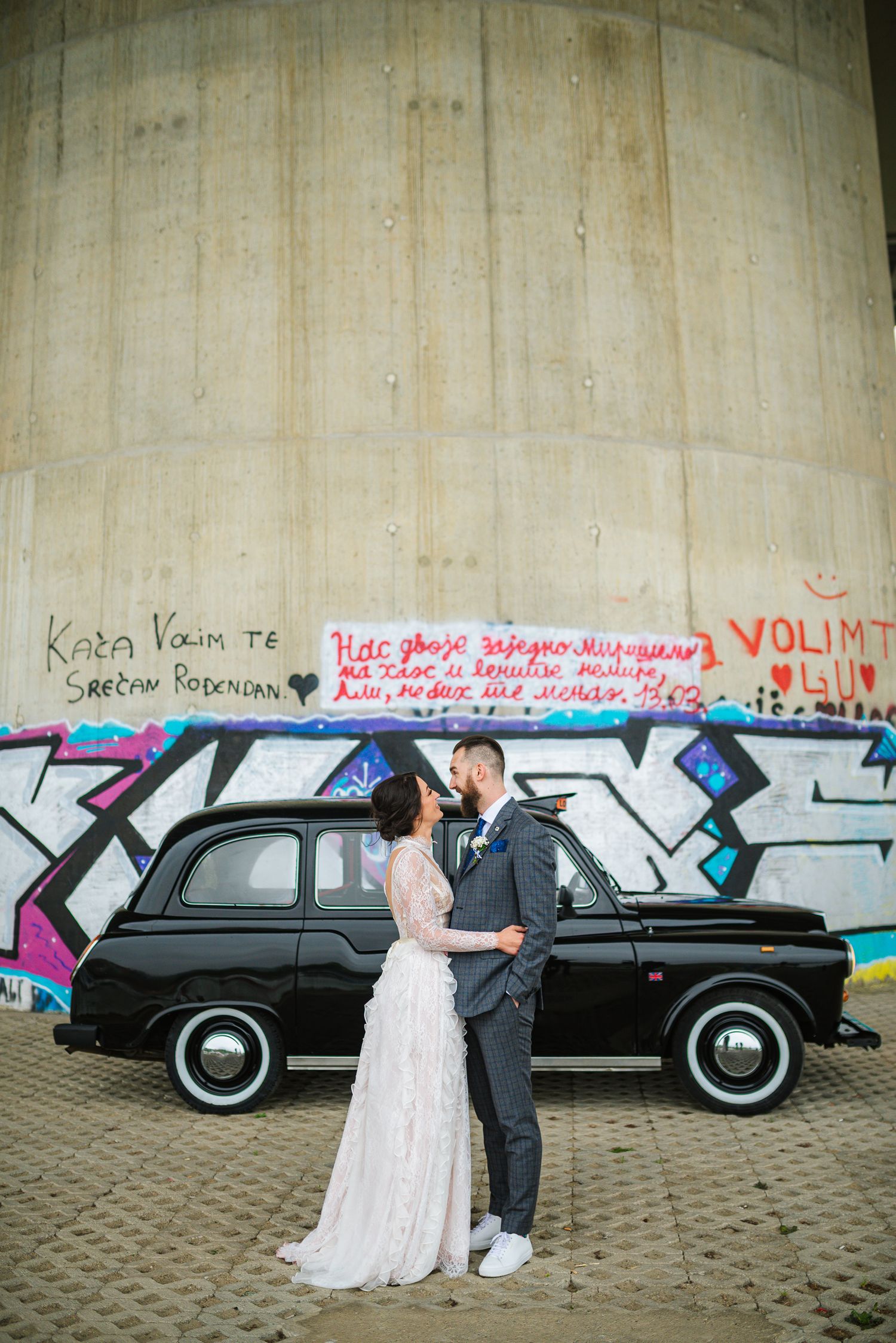 Bride and groom kissing in front of a black vintage taxi with graffiti-covered concrete wall backdrop.