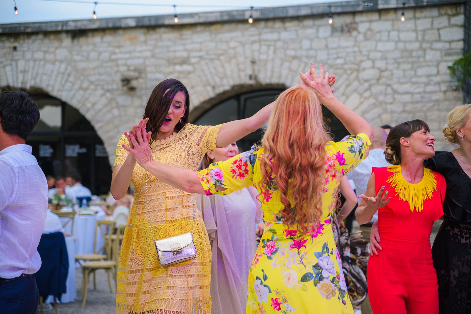 Women in colorful floral dresses dancing together at outdoor wedding reception.