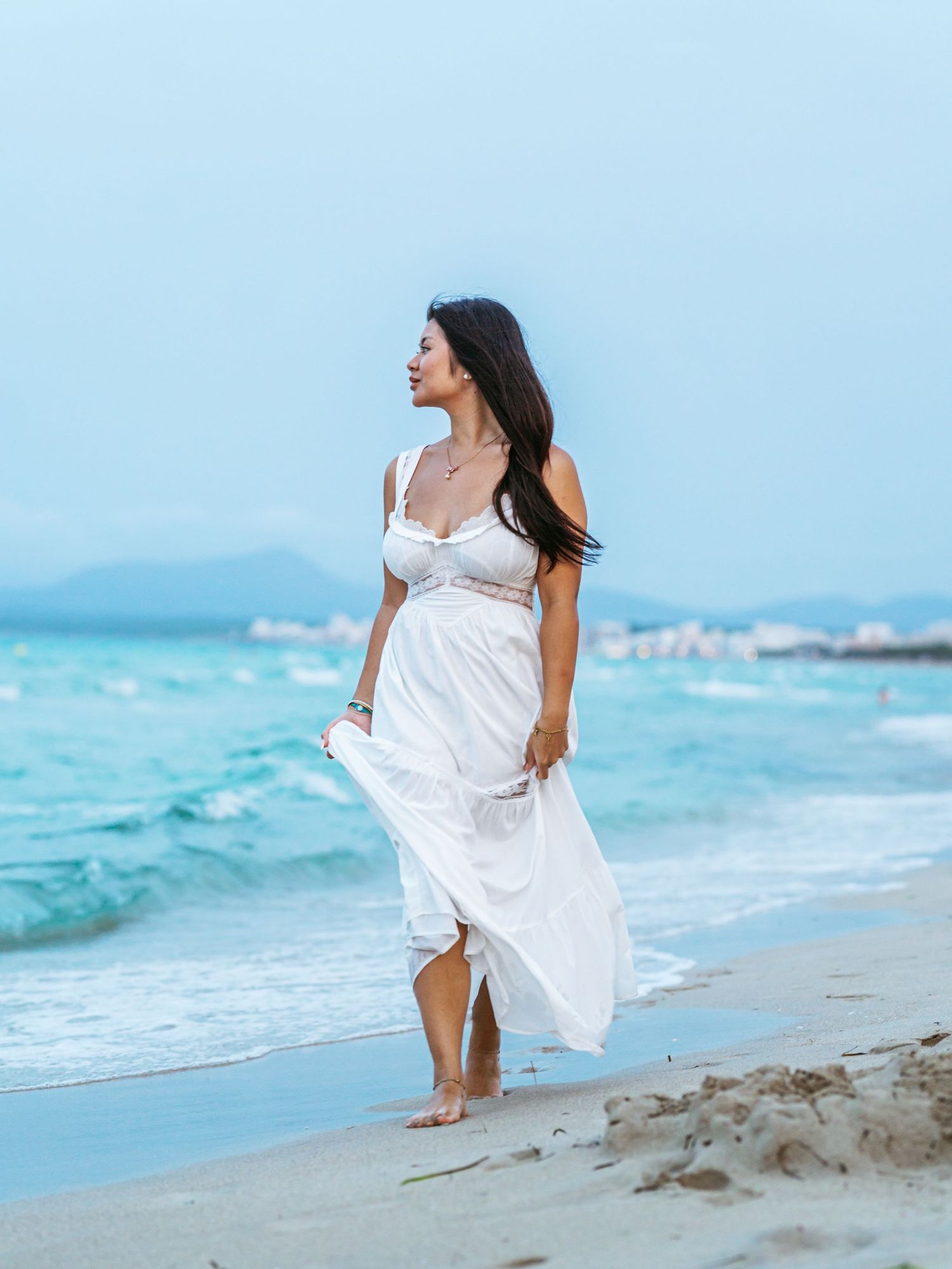 Woman walking along the beach in a white dress, natural lifestyle portrait photography by Herzenshaus Studio