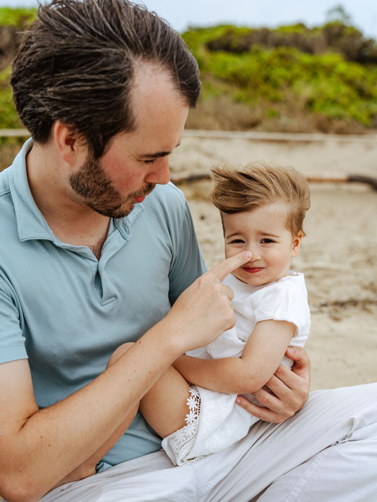 Father touching baby girl's nose while sitting on sandy beach.