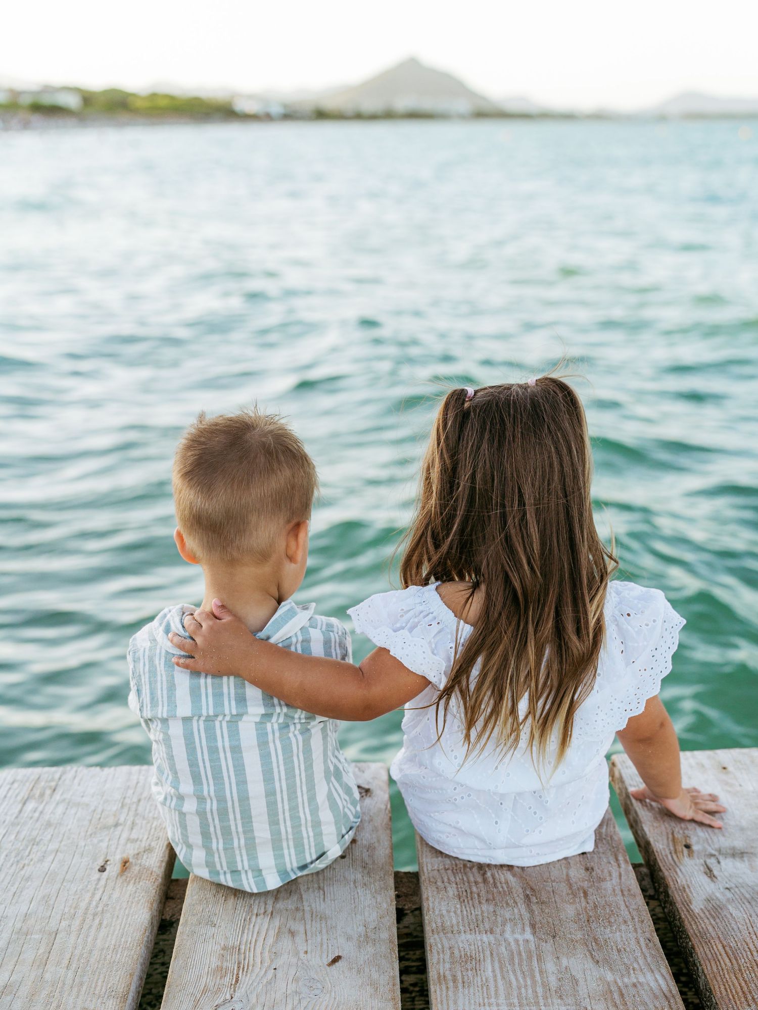 Two young siblings sit together on a wooden dock overlooking calm turquoise water.