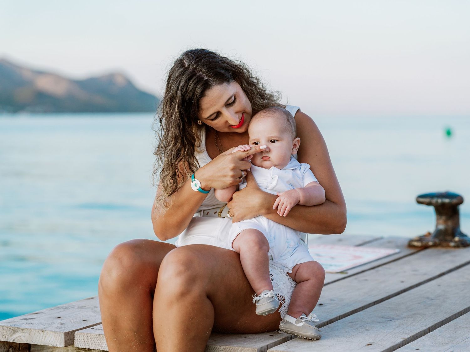 Young mother sitting on a wooden pier holding her baby while overlooking calm blue water.