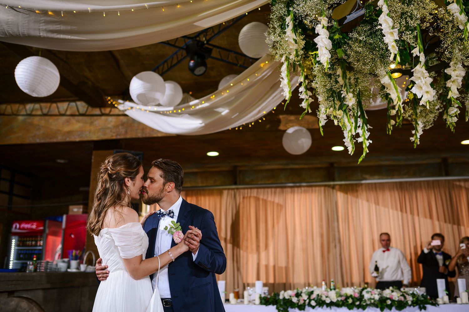 Bride and groom share a first kiss under floral ceiling decorations at an elegant wedding reception.