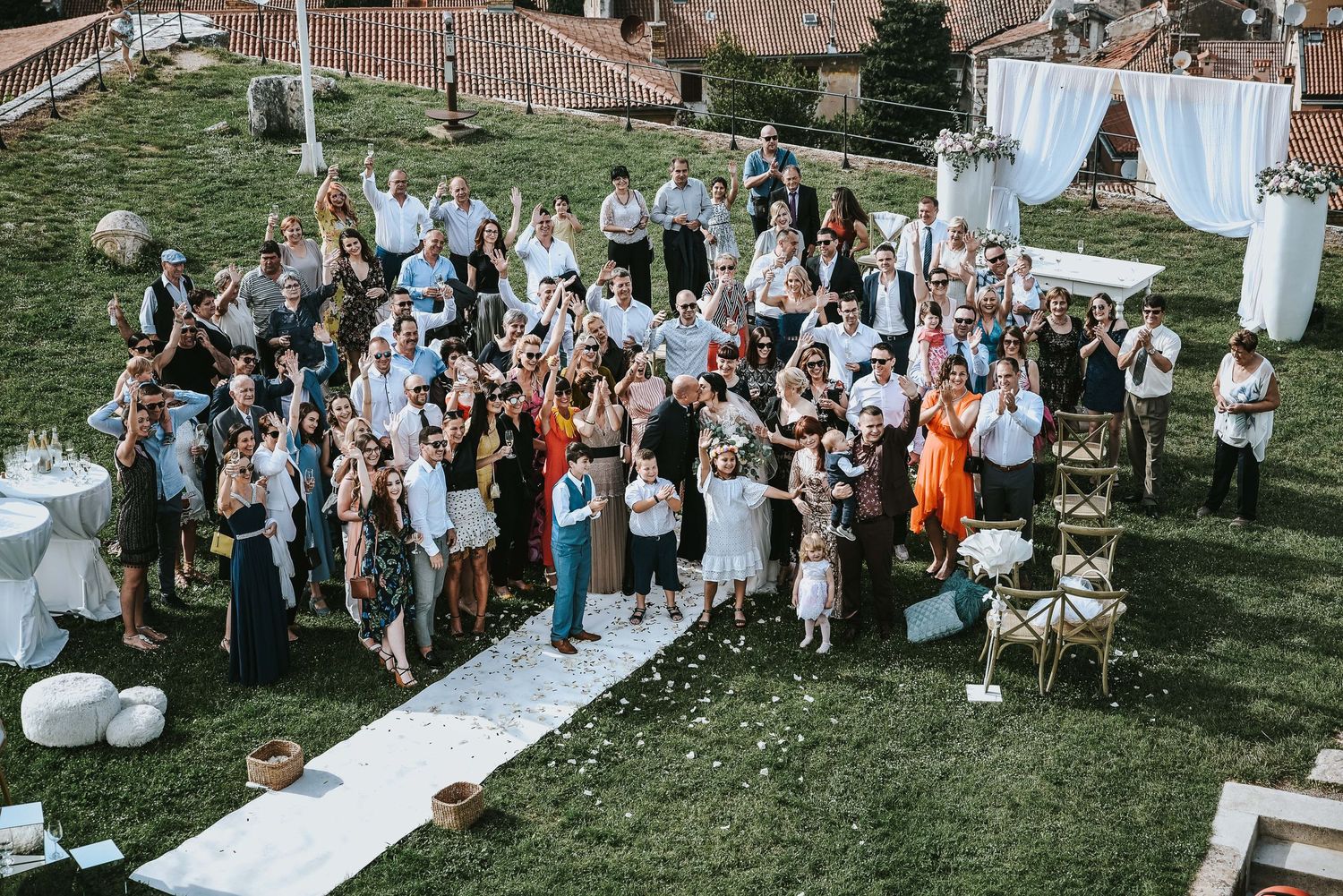 Aerial view of wedding guests gathered outdoors on a green lawn for a group celebration photo.