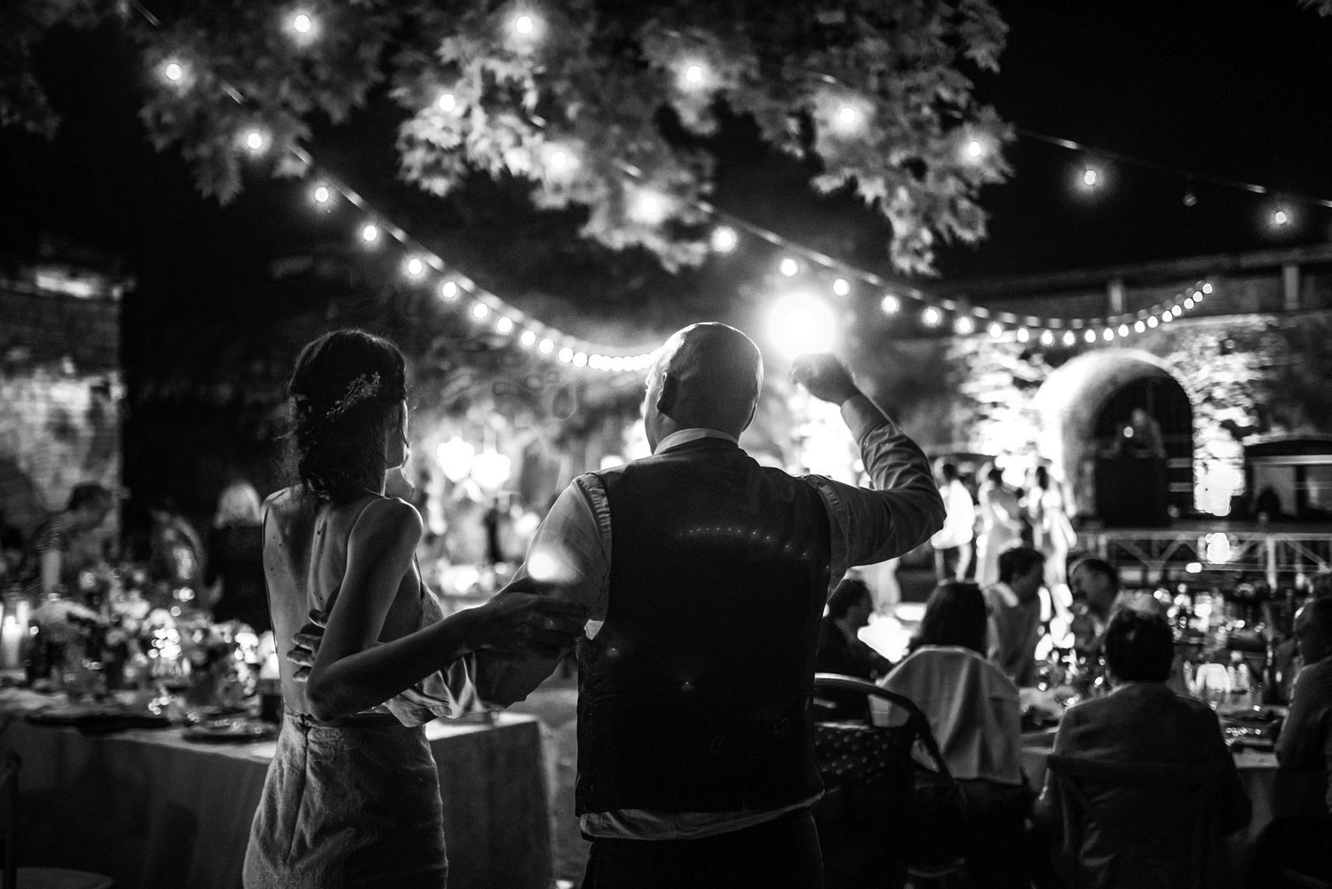 Black and white photo of wedding guests dancing under string lights at an elegant outdoor evening reception.