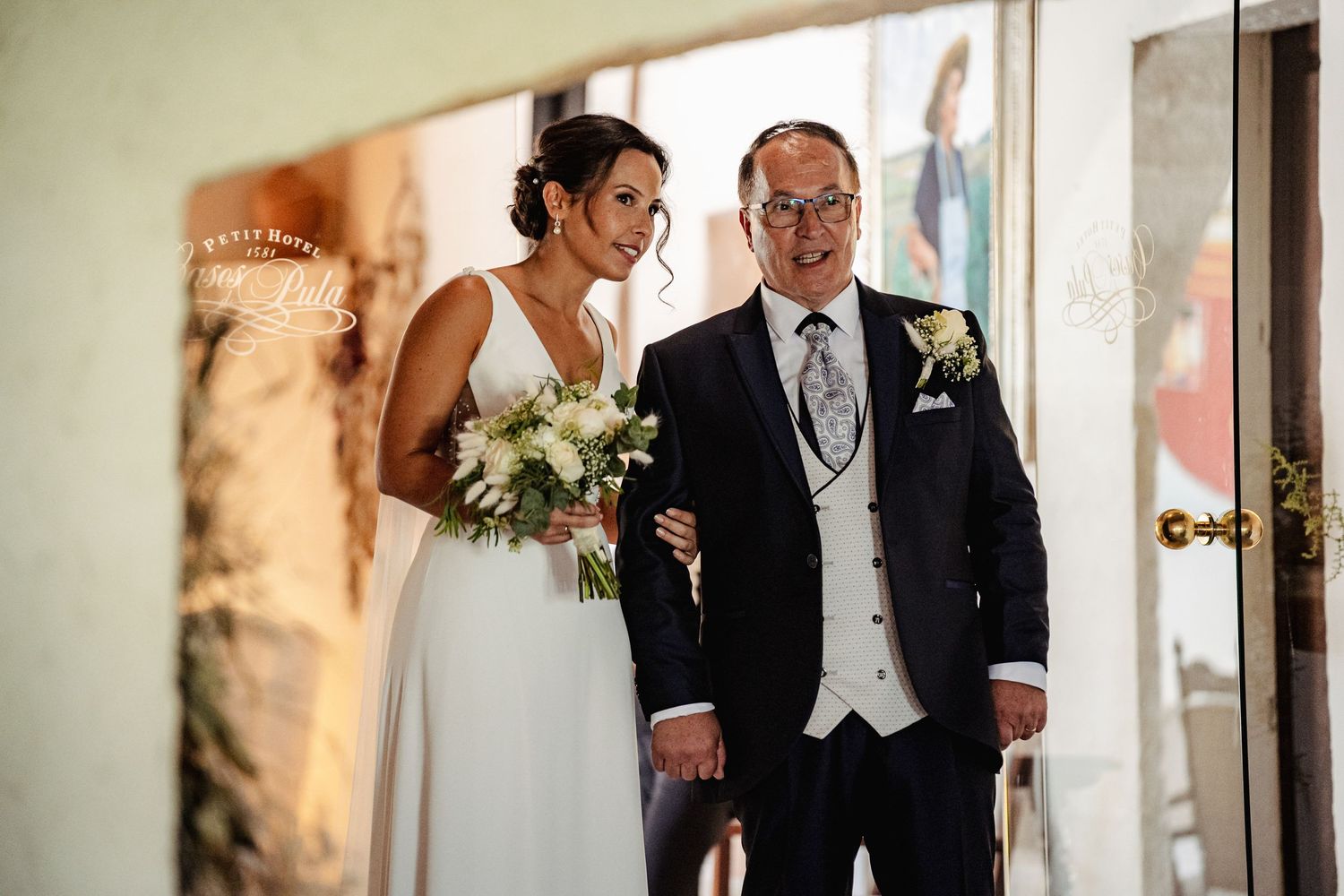 Bride holding white bouquet walking arm in arm with her father through a chapel doorway.
