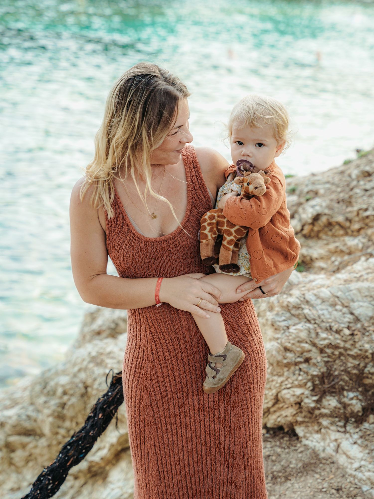 Young blonde mother in rust knit dress holding toddler with giraffe stuffed toy by rocky waterside.