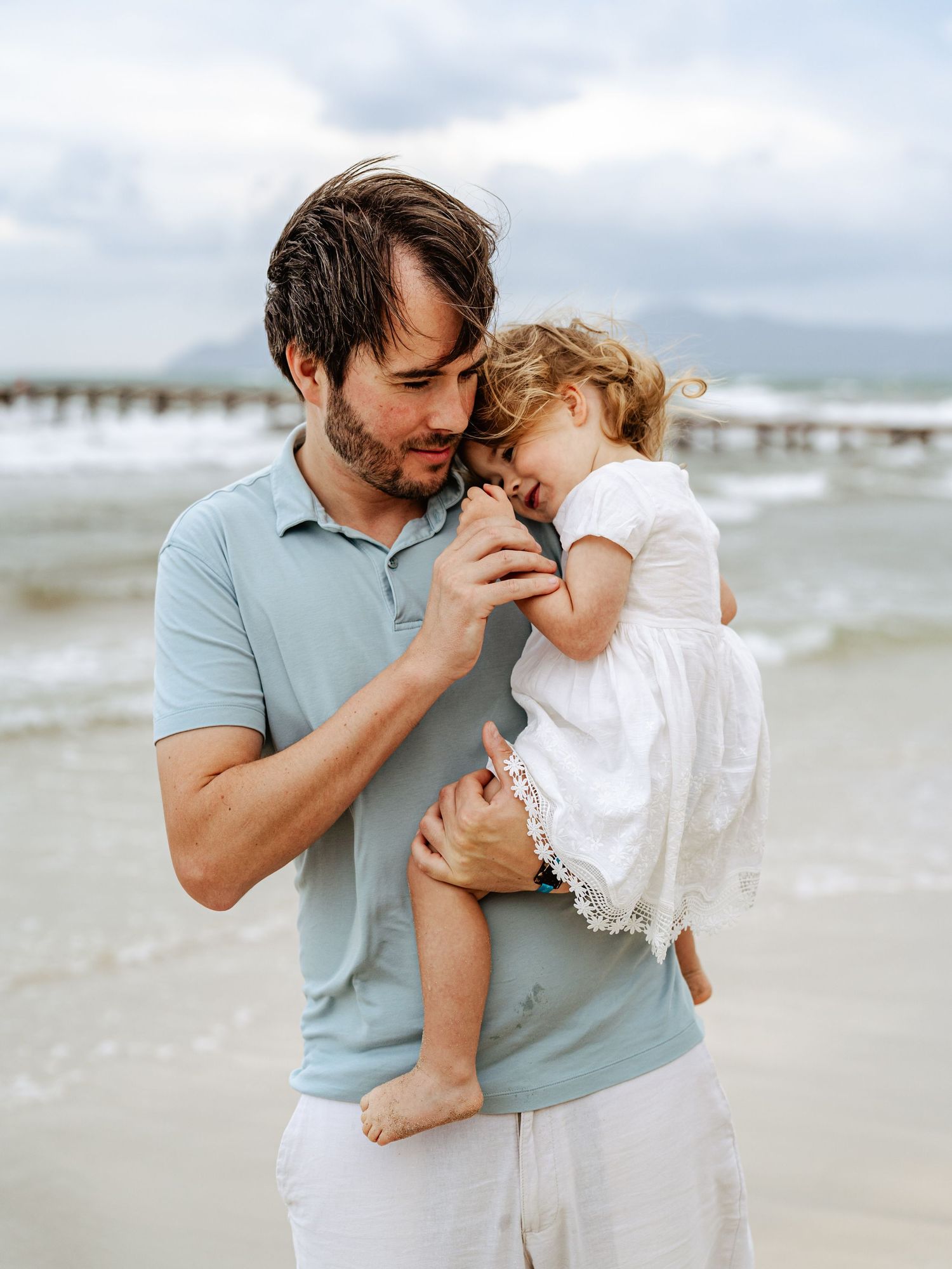 Father holding toddler close on a windy beach.