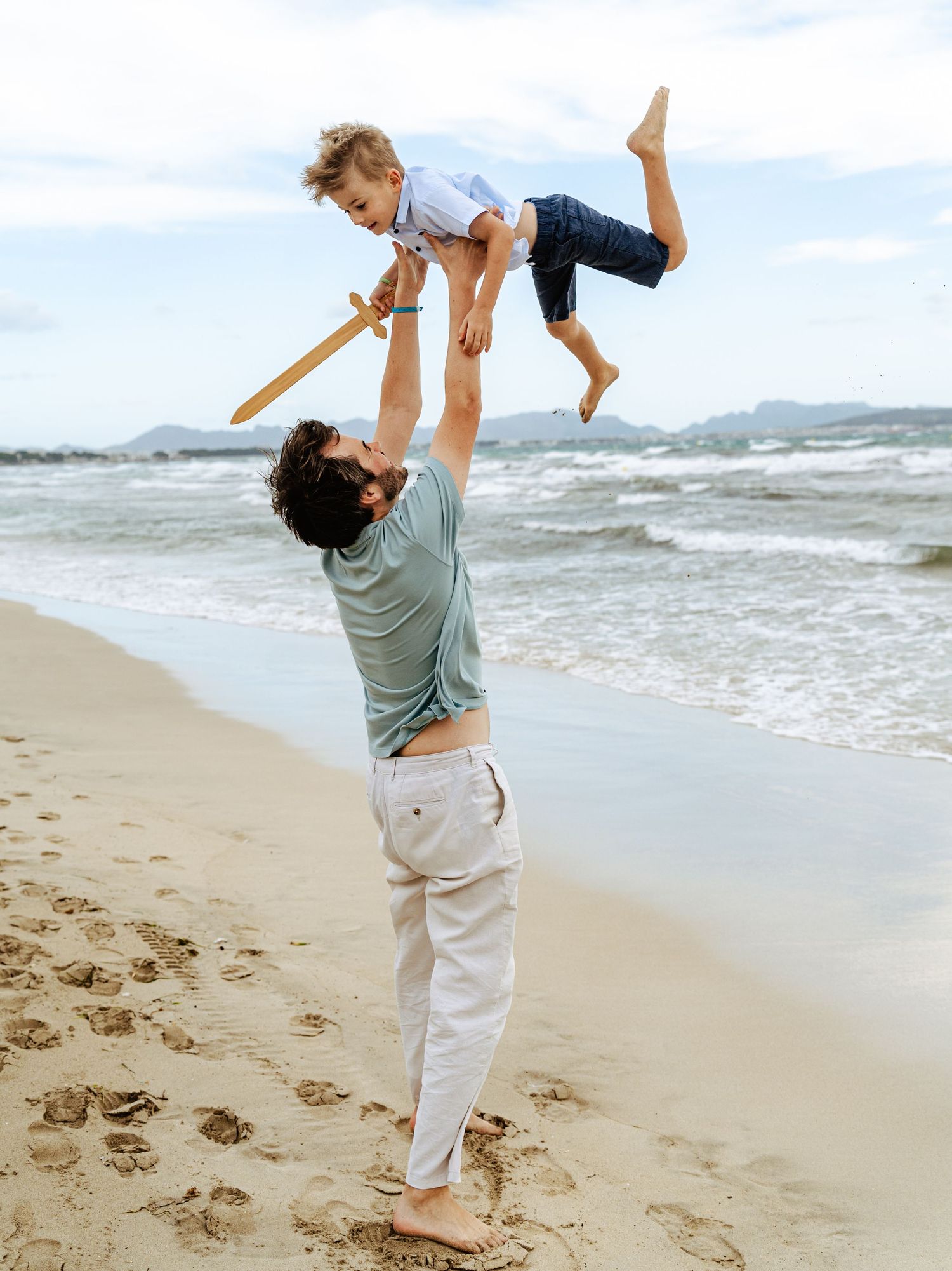 Father lifting young boy with wooden sword high into the air on a sandy beach by the ocean.