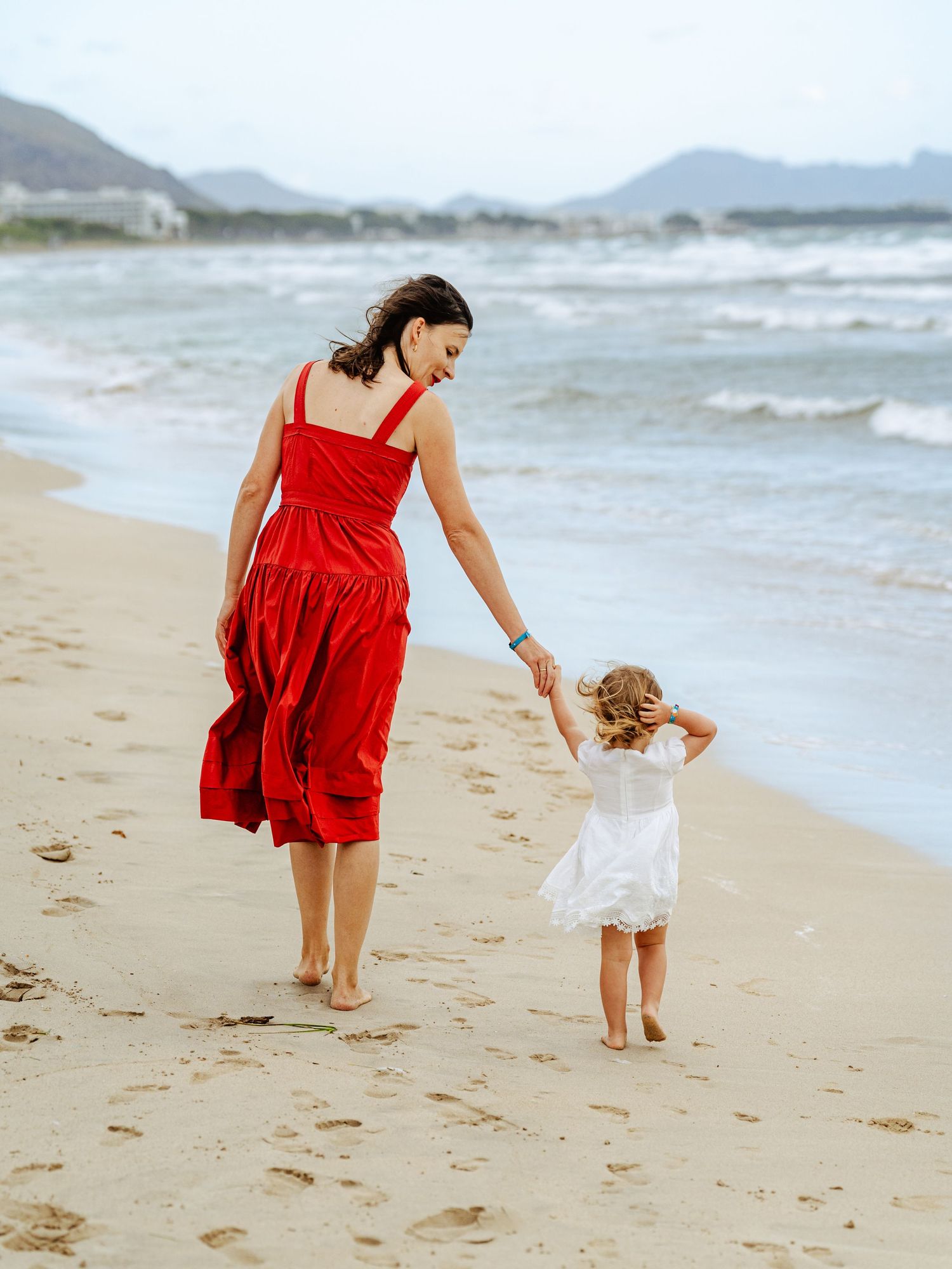 Mother in red dress holds toddler girls hand walking along sandy beach.