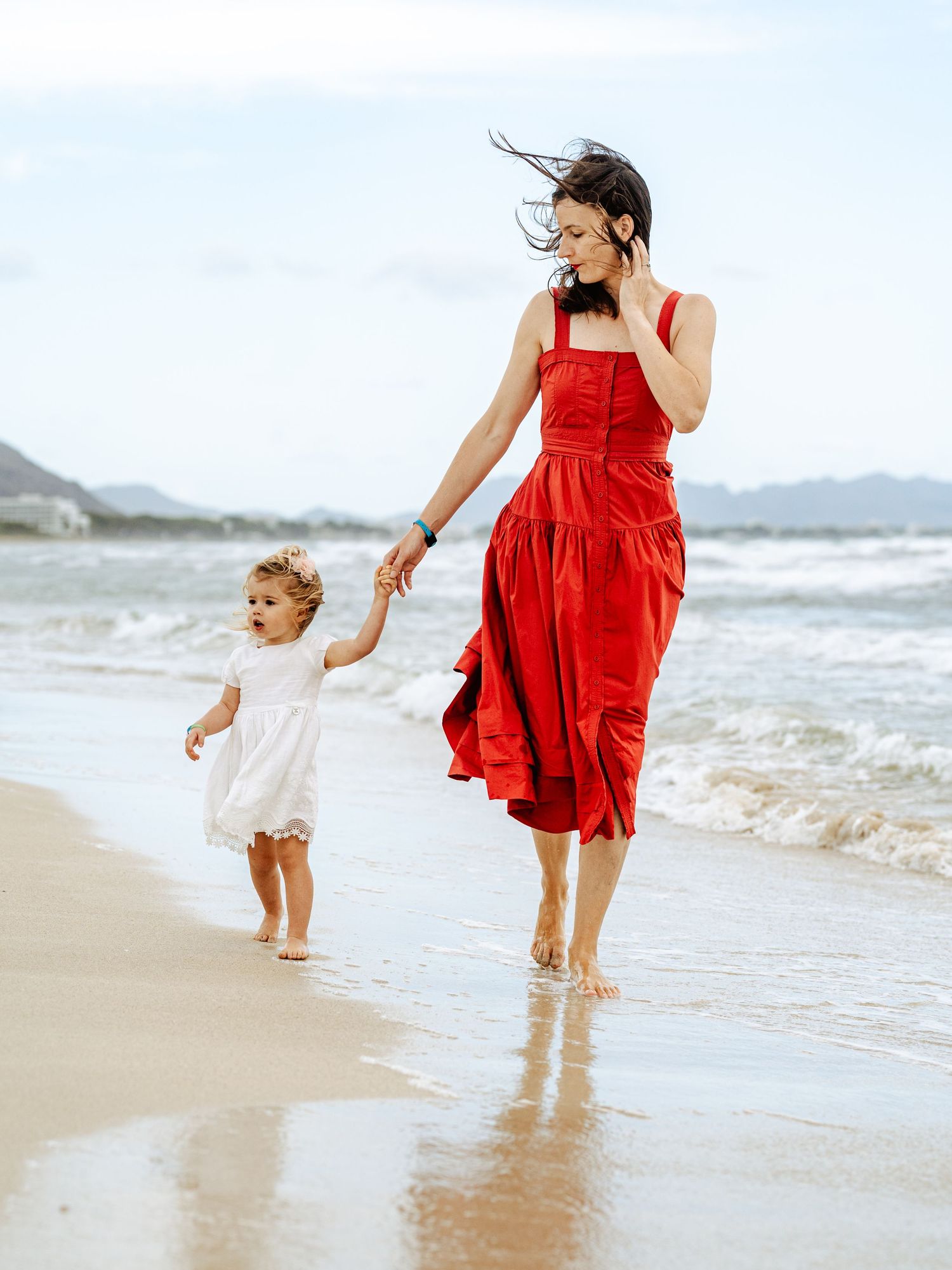 Smiling mother in red dress holds her toddler's hand while strolling along a windy beach.