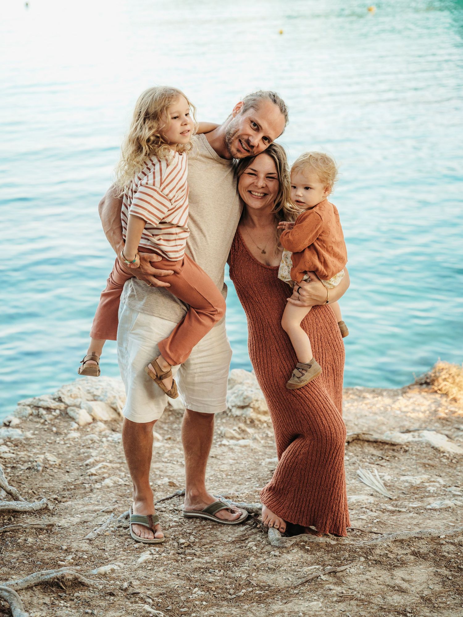 Happy family of four posing together on rocky shore with blue ocean backdrop.