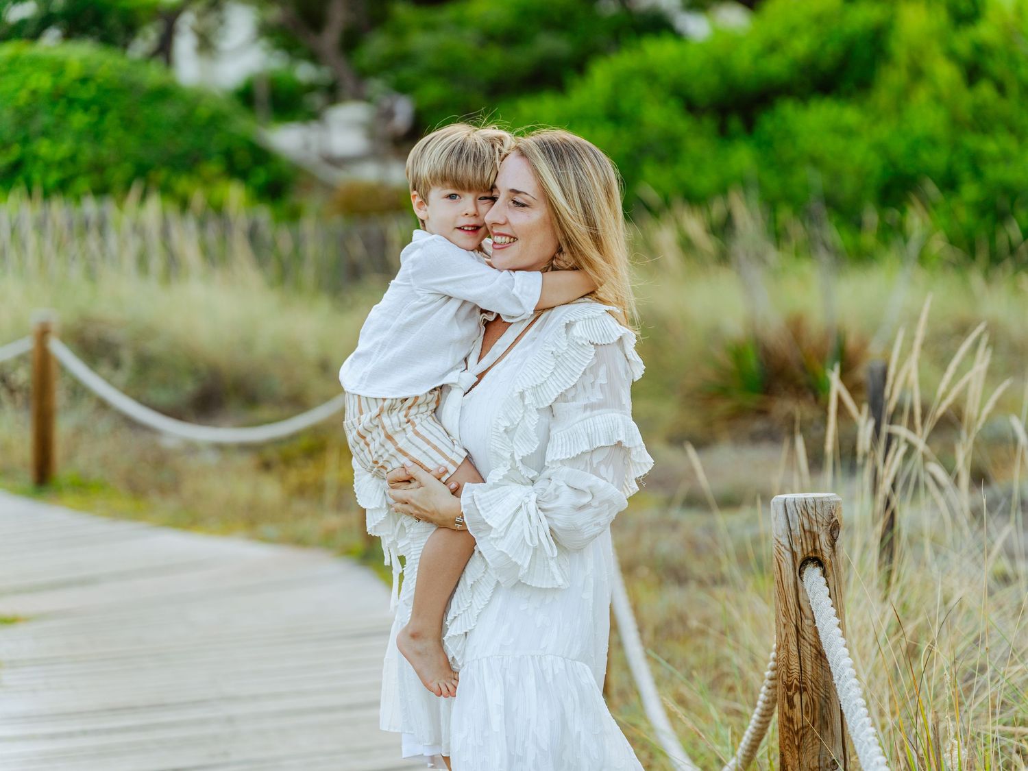 Loving mother in white ruffled dress hugging her young son on a sunny boardwalk.