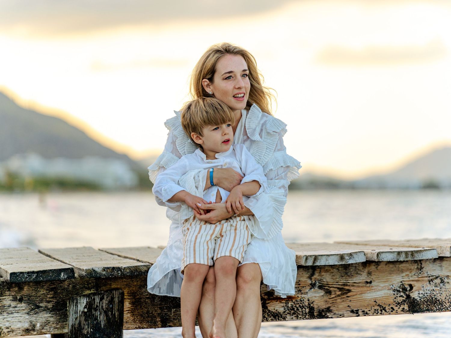 Mother hugging young son while sitting on beach pier at sunset.