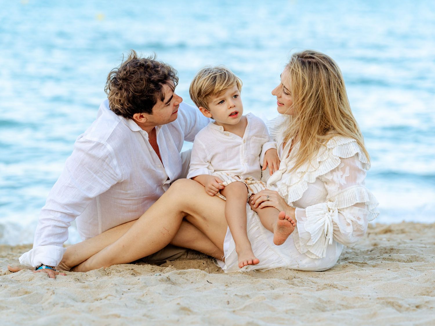 Happy family of three sitting on sandy beach with parents lovingly looking at their young son.