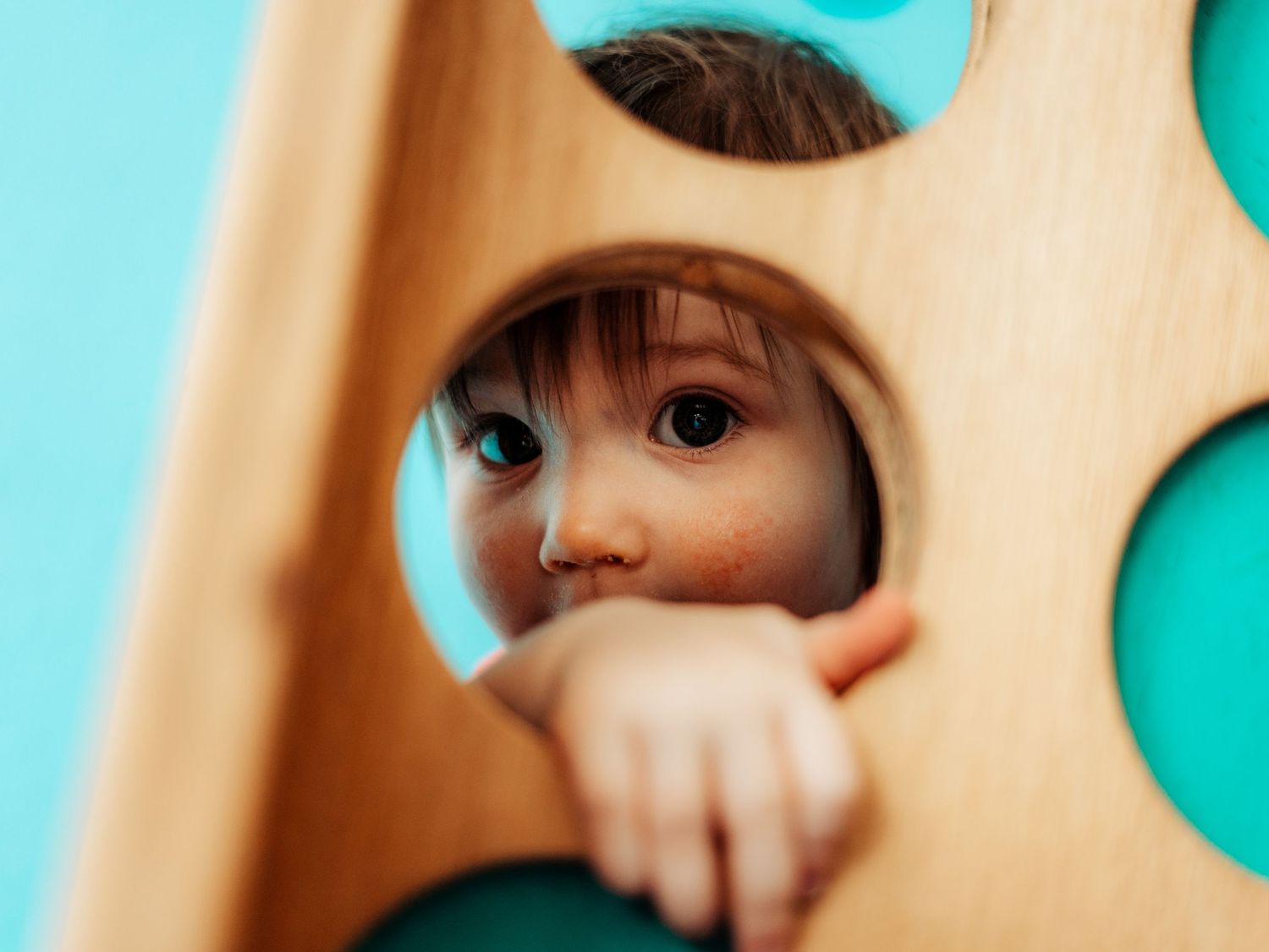 Playful toddler girl peeking through holes in wooden play structure against bright teal background.