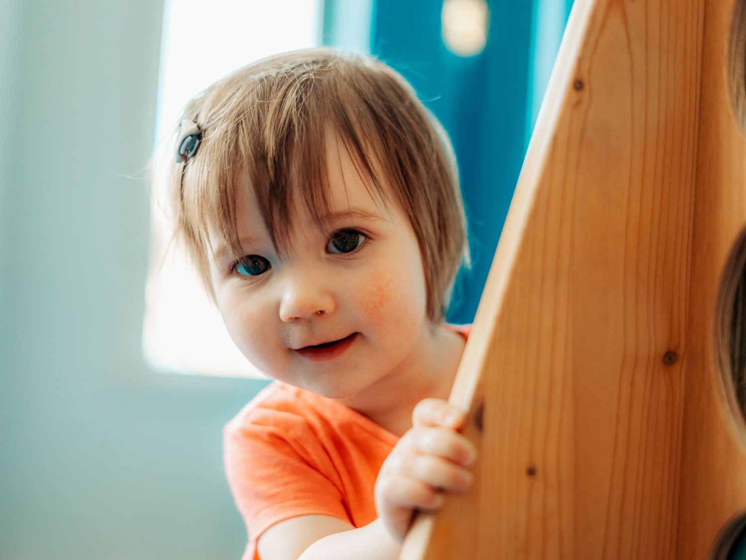 Happy toddler girl in orange shirt smiling while holding onto a wooden play structure indoors.