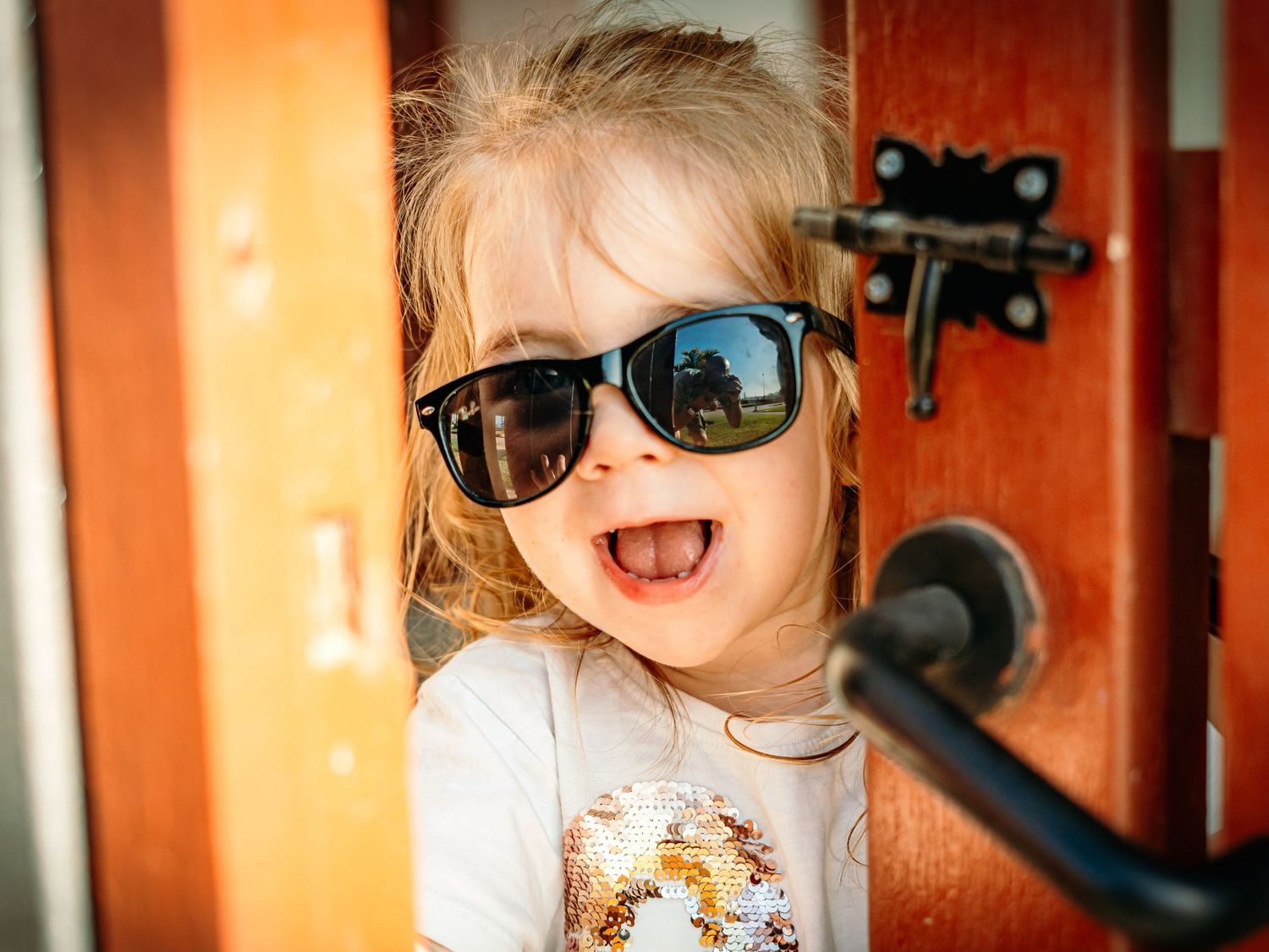 Adorable blonde toddler girl laughing while wearing black sunglasses at a wooden door.