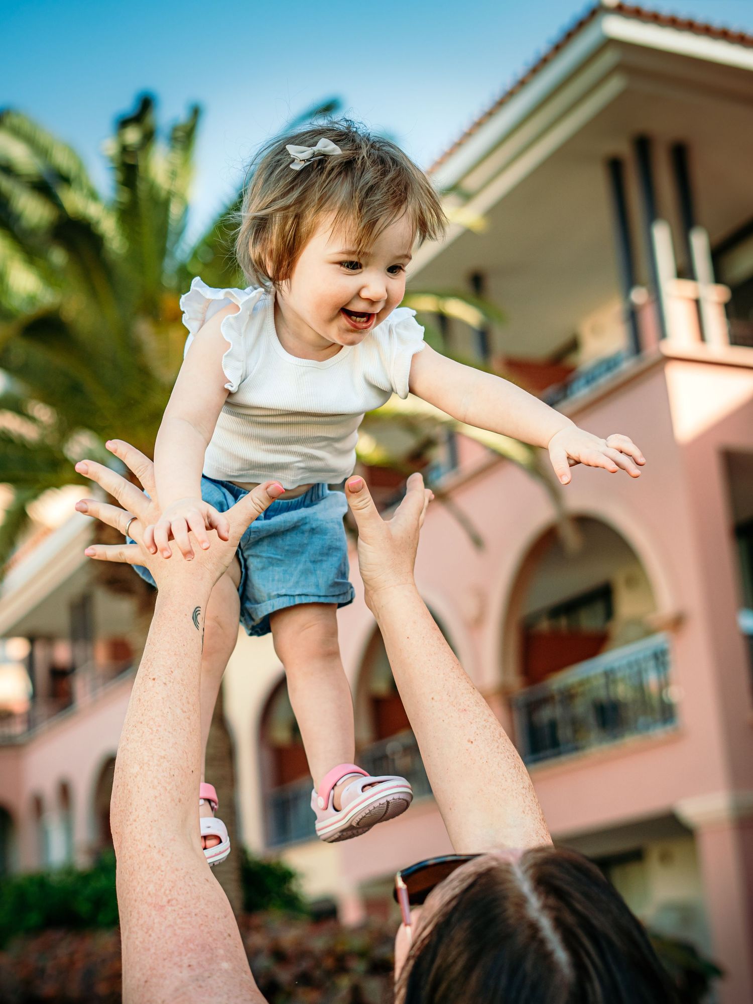 Smiling toddler girl tossed in the air by a parent outside a pink resort with palm trees.