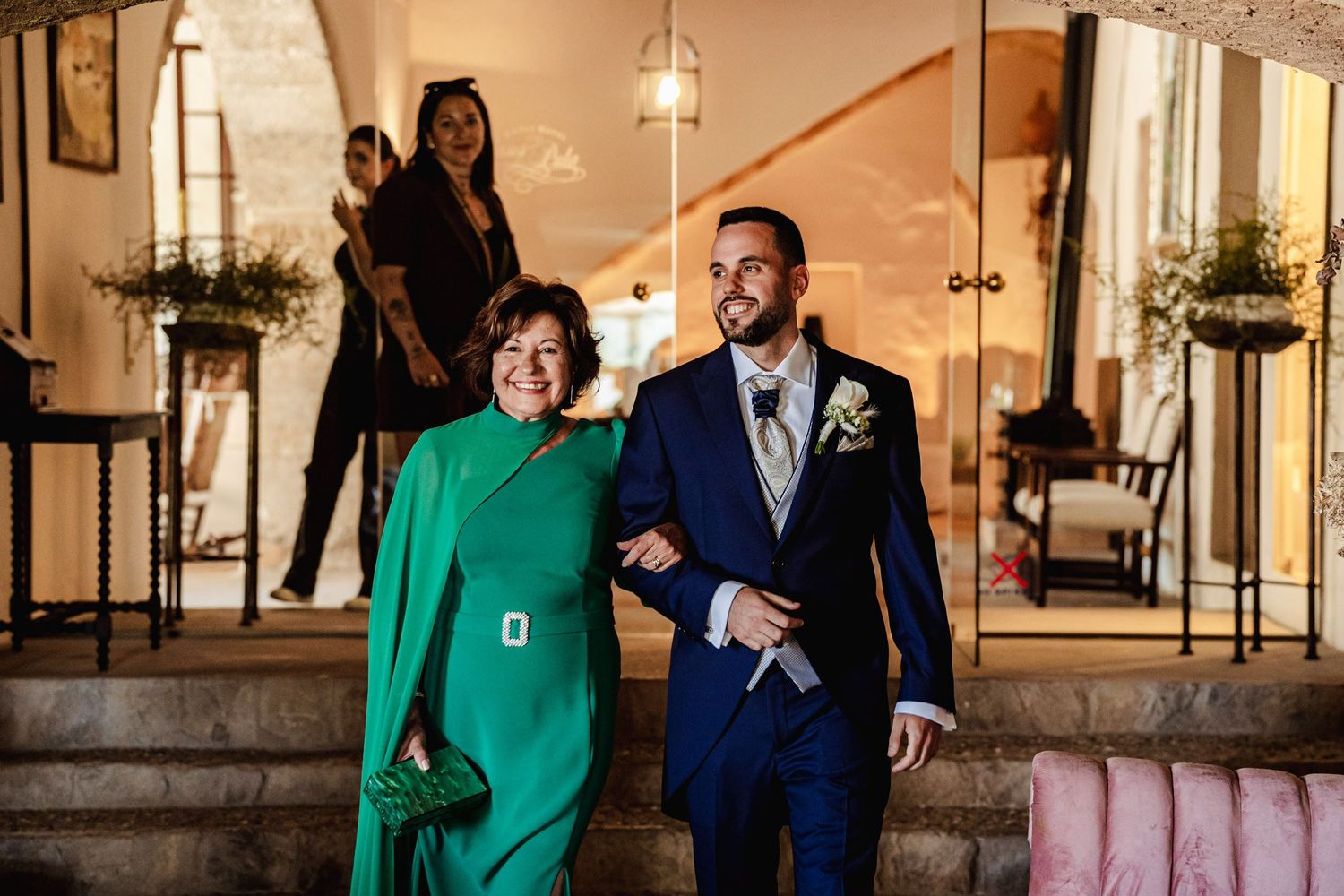 Groom in navy suit walks with woman in green dress through elegant wedding venue entrance.