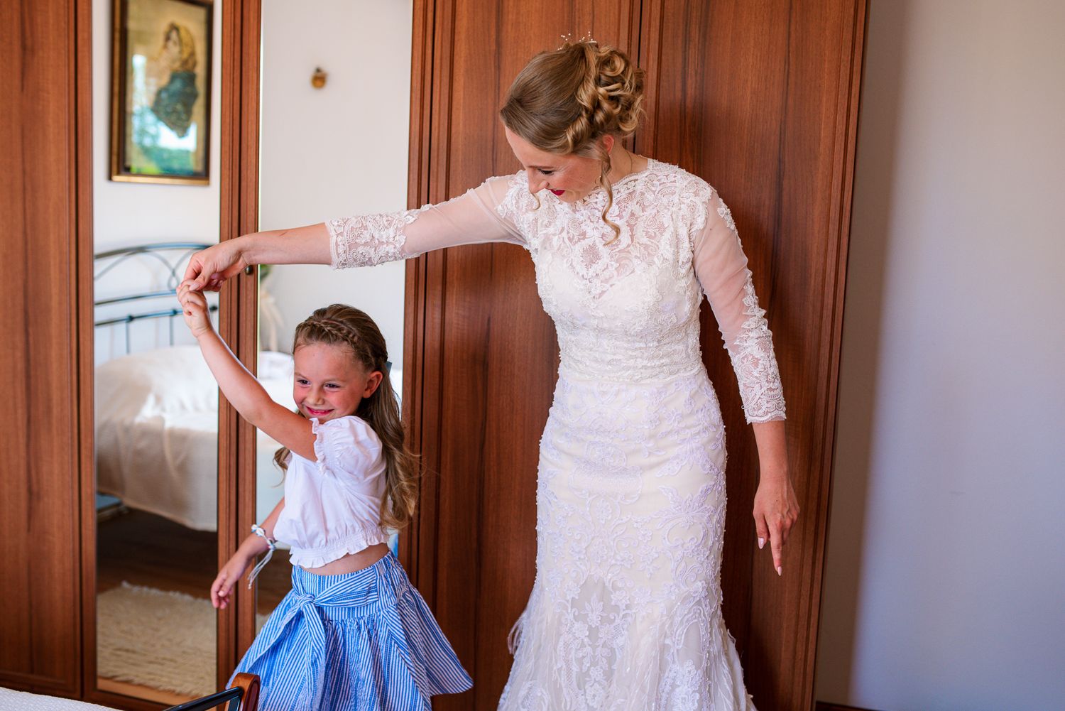 Bride in elegant lace wedding dress dancing with young flower girl in blue skirt near mirror.