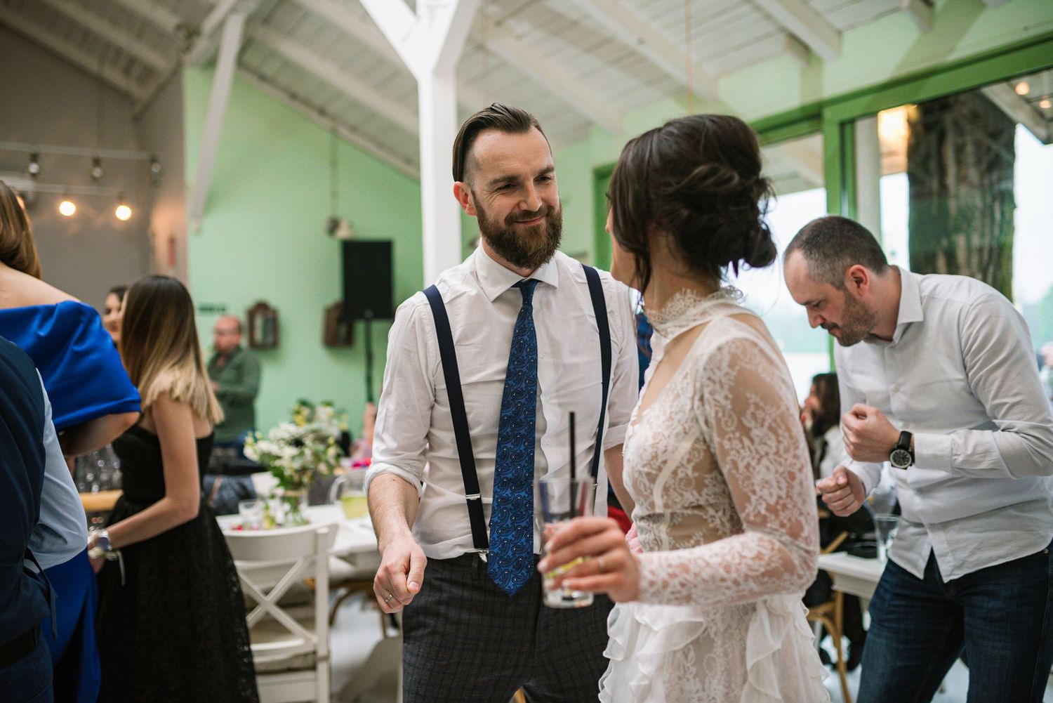 Groom in suspenders and tie chats with bride in lace dress at wedding reception.