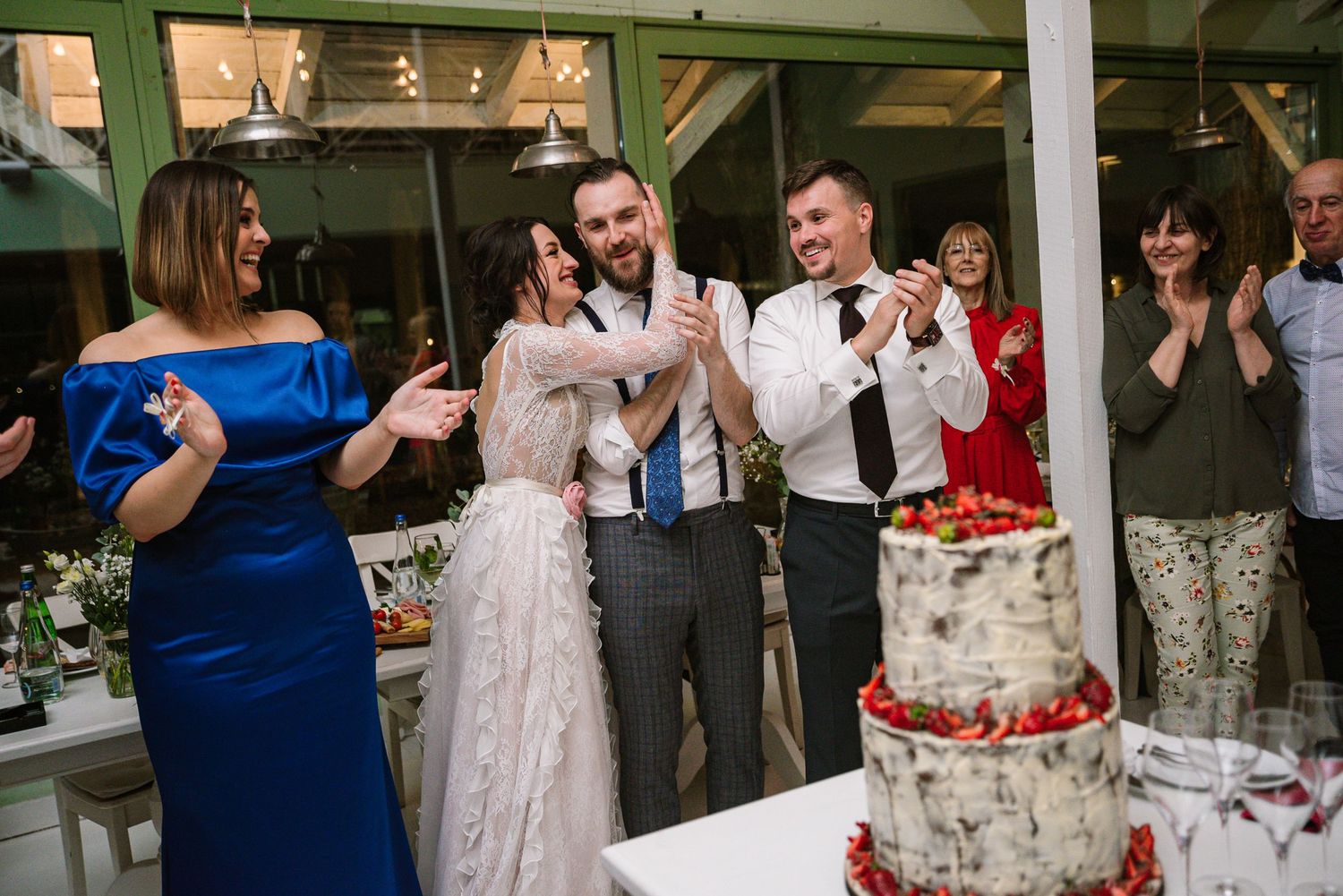 Bride and groom celebration beside their wedding cake as family and friends applaud during the reception.