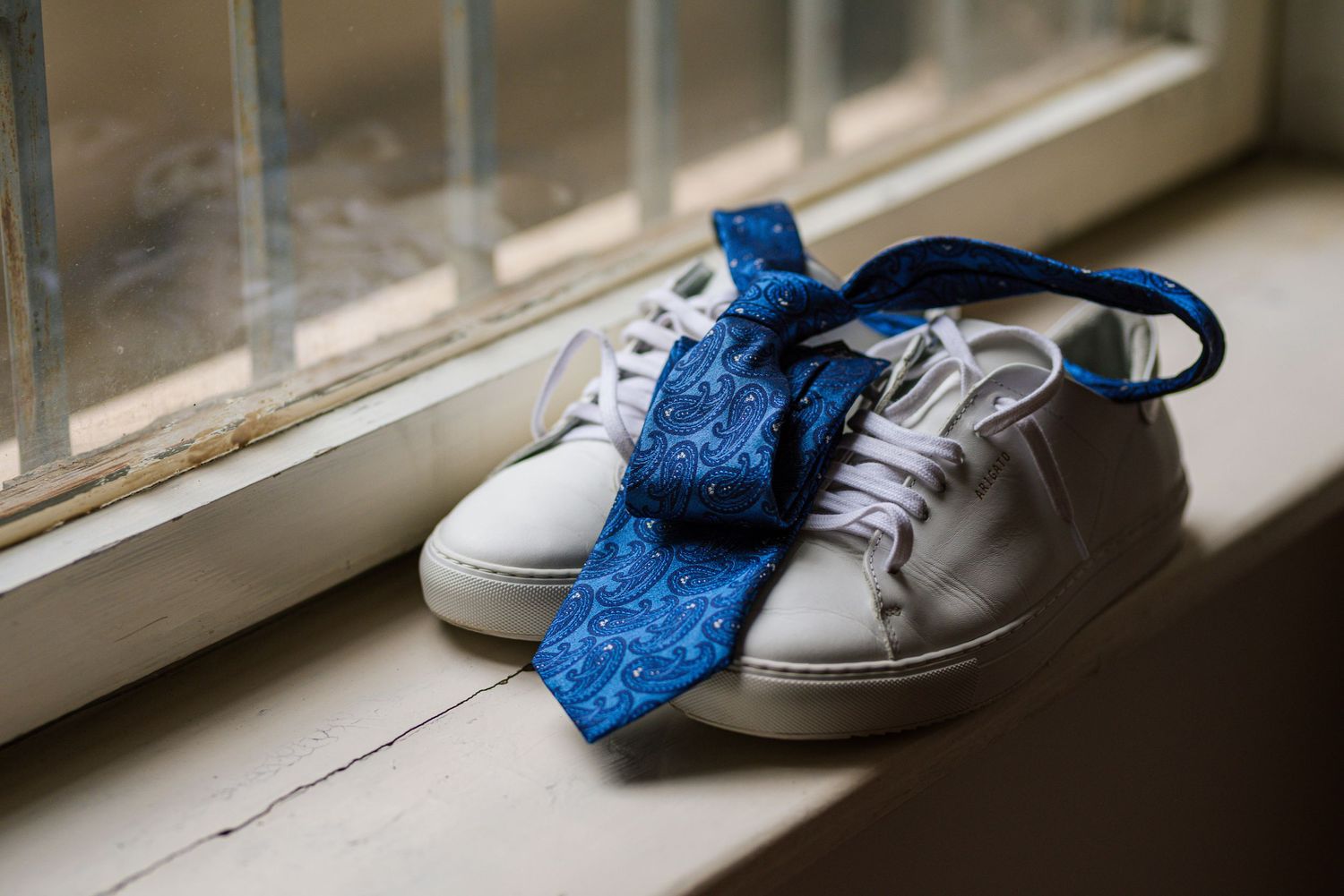 White sneakers with a blue patterned tie draped over them on a windowsill.