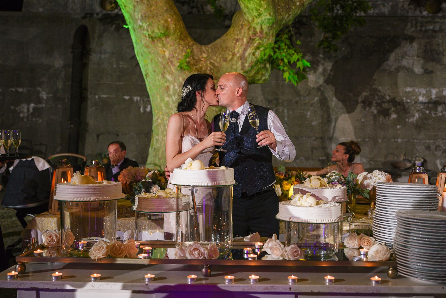 Bride and groom share a romantic kiss over champagne glasses at their candlelit outdoor wedding reception.